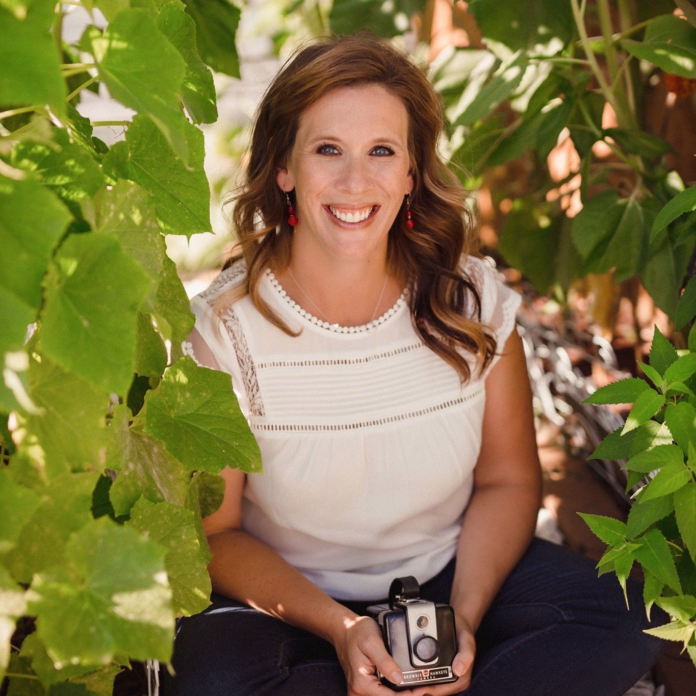 A woman with long, wavy brown hair, smiling, sitting outdoors among green leaves, holding a vintage camera.