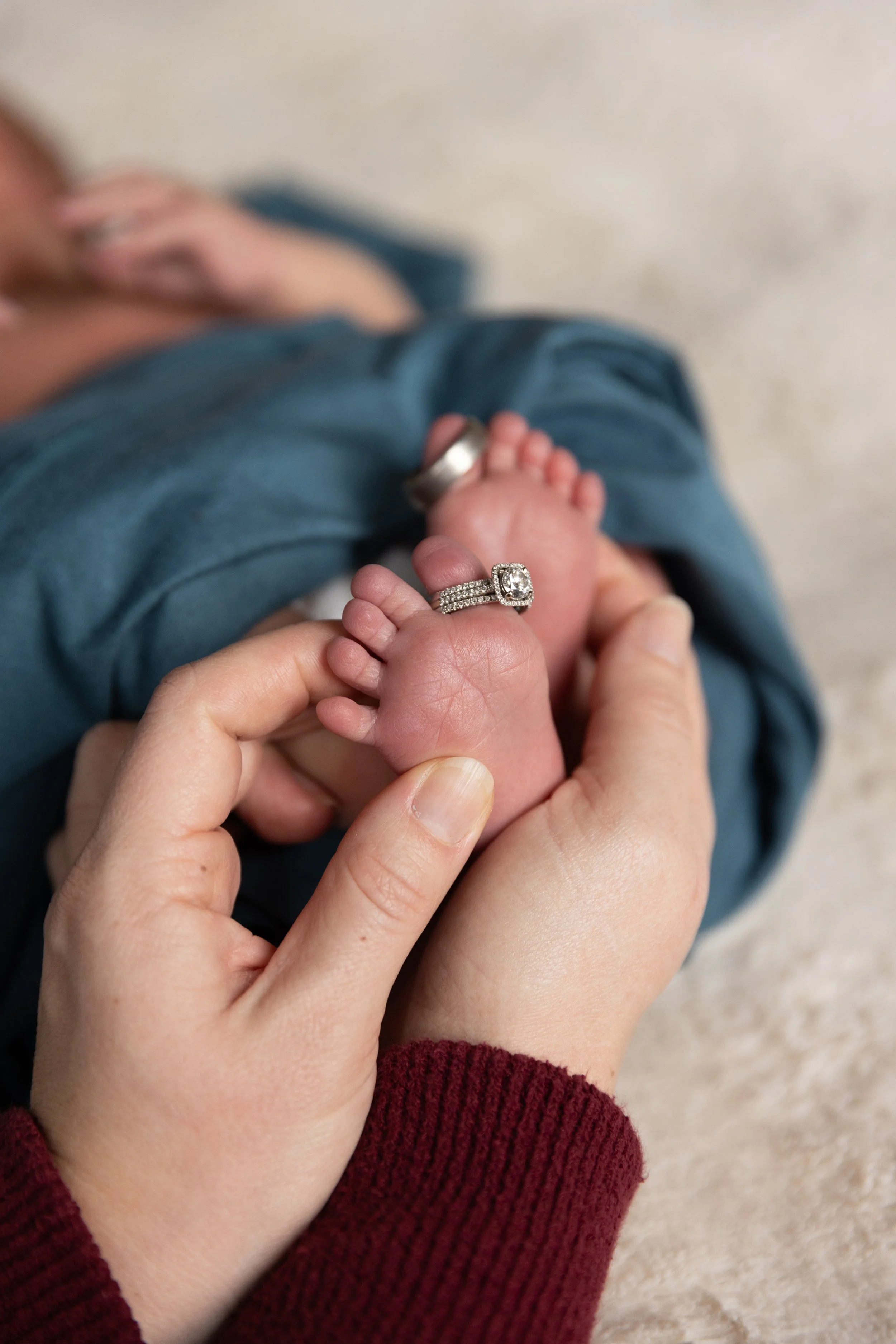 A close-up of a person holding a tiny baby’s foot, which has a diamond ring on the toe. The adult's hand supports the baby's foot, and the background shows the adult wearing a red sweater and jeans.