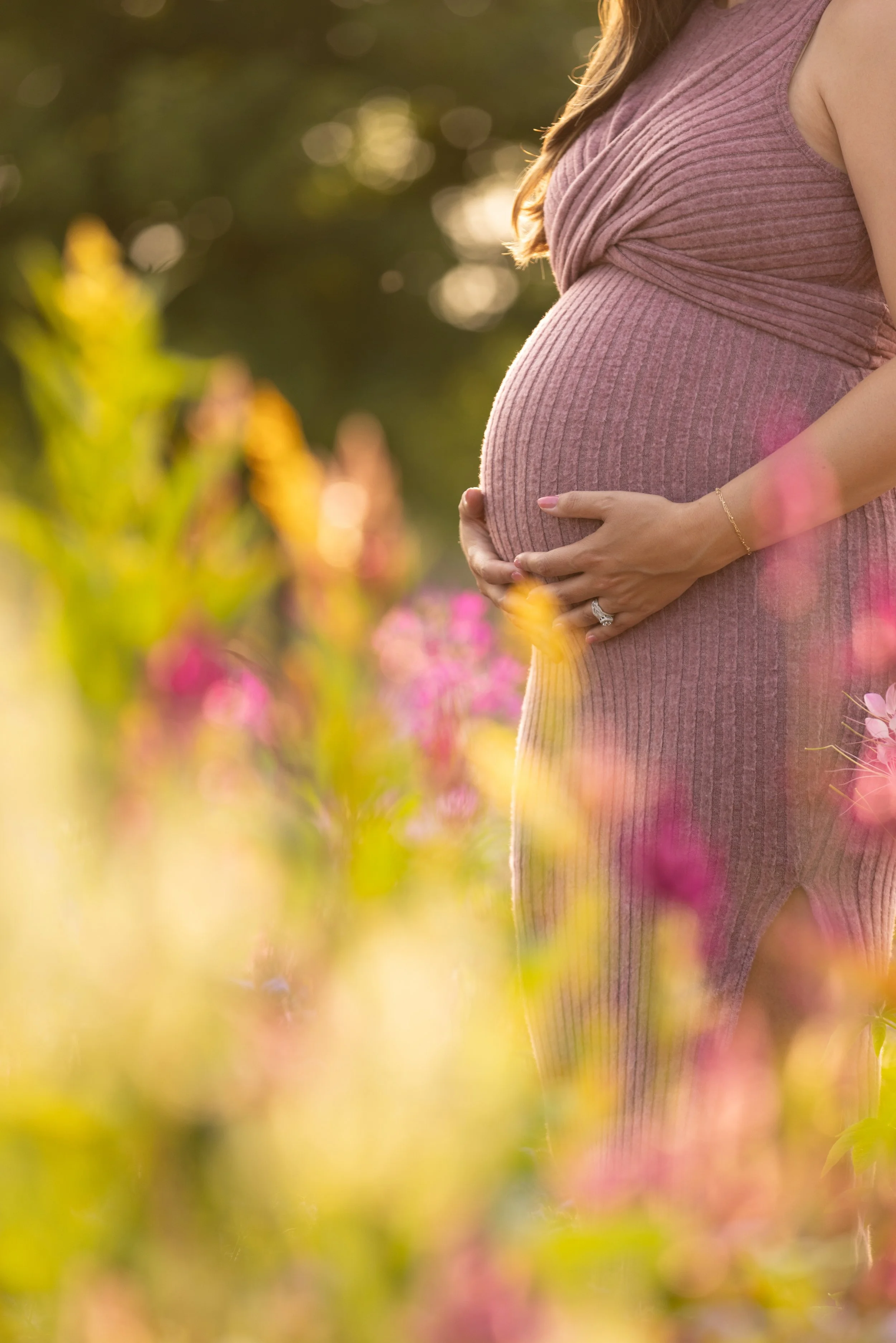 A pregnant woman in a pink dress is standing outdoors, holding her belly with both hands. The background features pink flowers and greenery with sunlight filtering through.