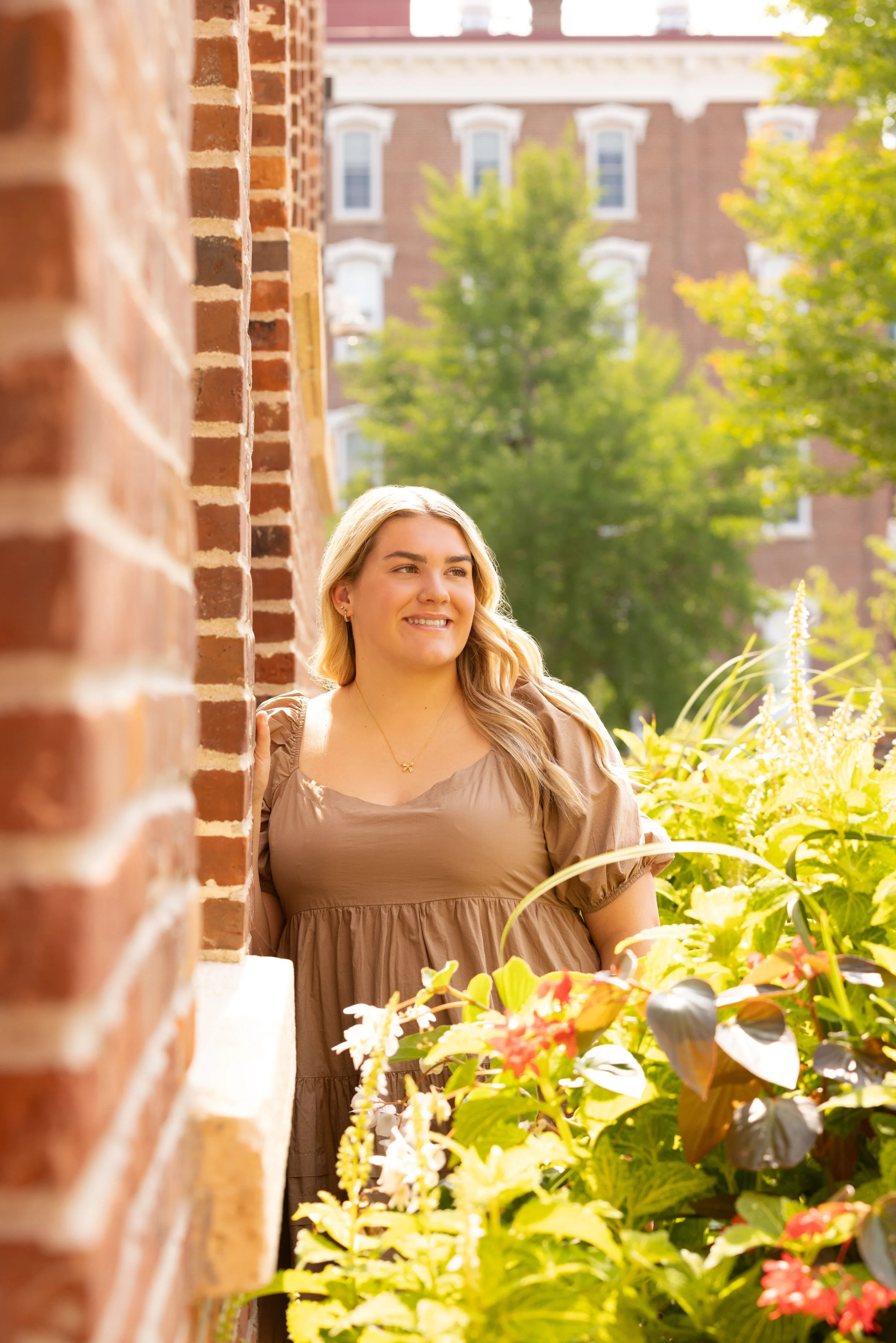 A young woman standing on a balcony surrounded by green plants and flowers, smiling with her head turned slightly to the side, with a brick building and trees in the background on a sunny day.
