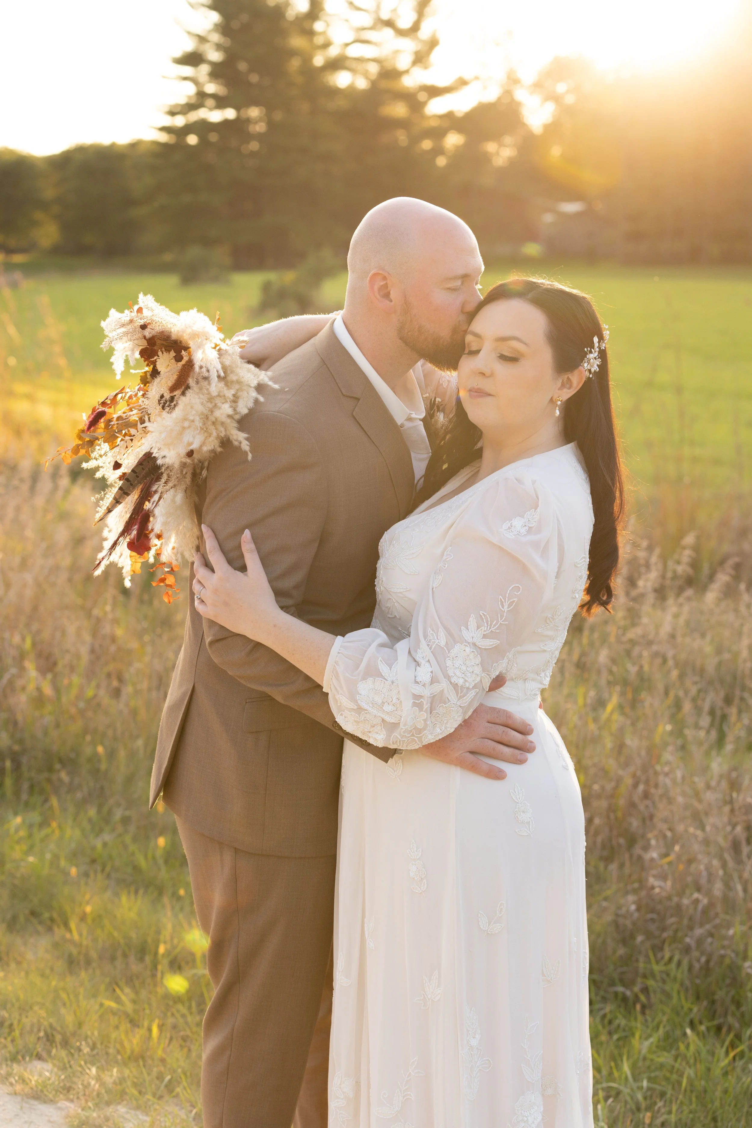 A wedding couple embracing outdoors during sunset, with the groom kissing the bride on the forehead. The bride is wearing a white lace dress with floral embroidery, and the groom is dressed in a brown suit. They are in a grassy field with trees in th
