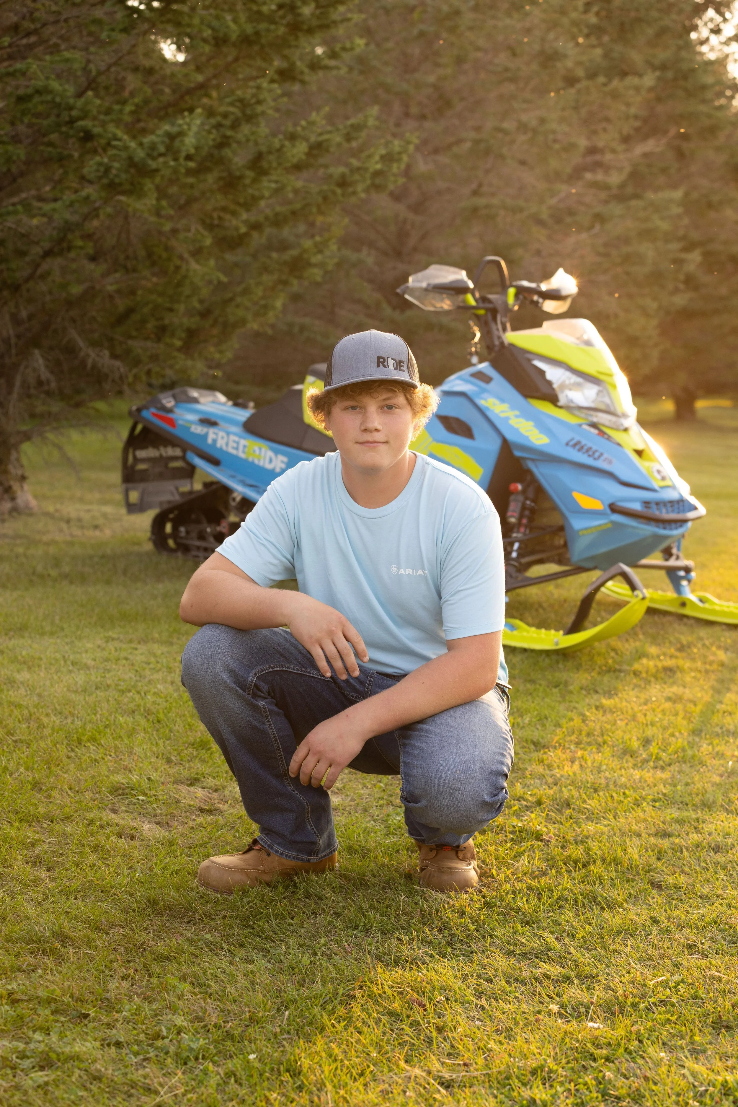 A young man crouching on grass shirtless in a blue T-shirt, with a snowmobile in the background during sunset.