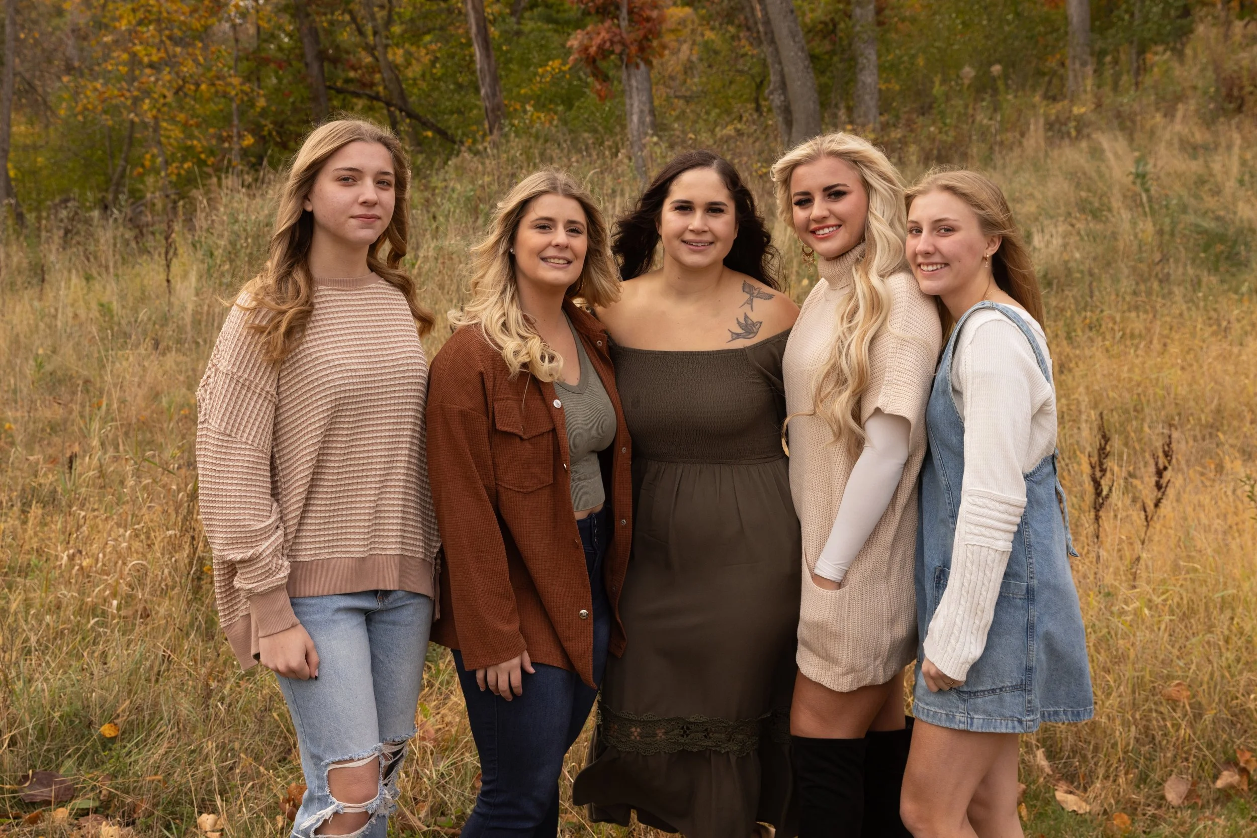 Five young women standing together outdoors in a grassy, fall landscape with trees and autumn leaves, smiling at the camera.