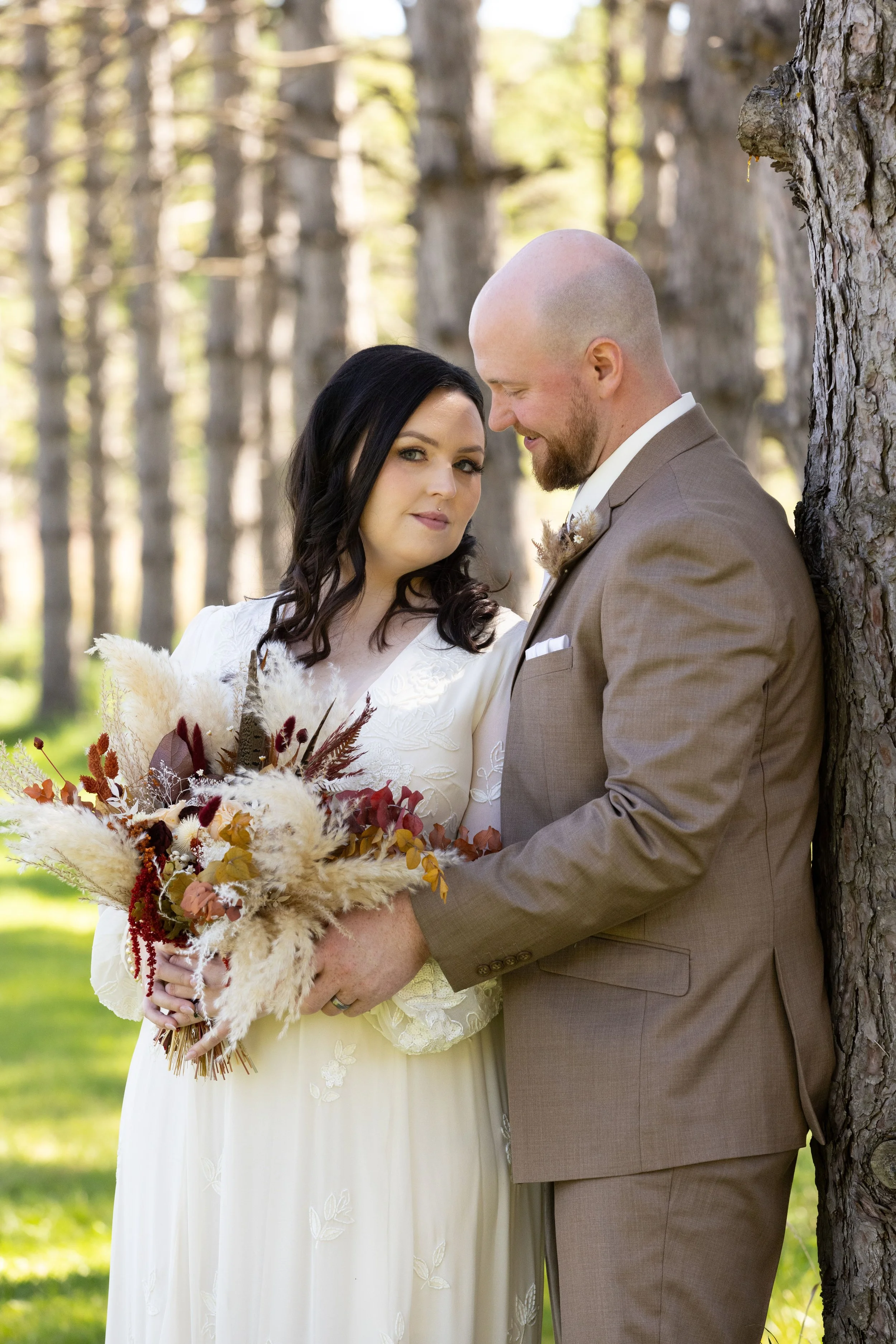 A bride and groom stand close together in a wooded outdoor setting, with the bride holding a bouquet of autumn-colored flowers and grasses, and the groom smiling with his head slightly tilted toward her.