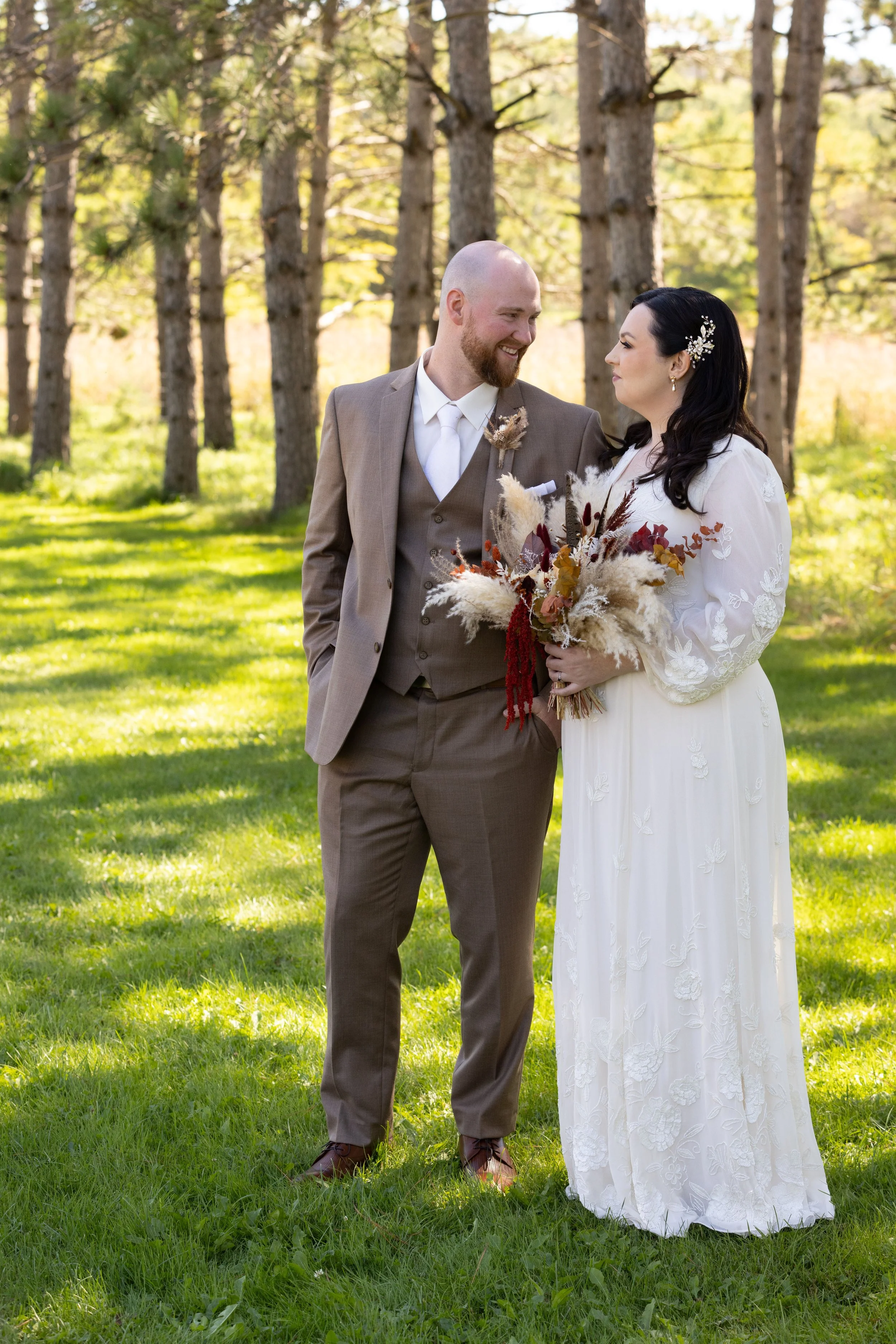 A groom and bride standing outdoors in a grassy area with trees, gazing at each other, with the bride holding a bouquet of dried flowers.
