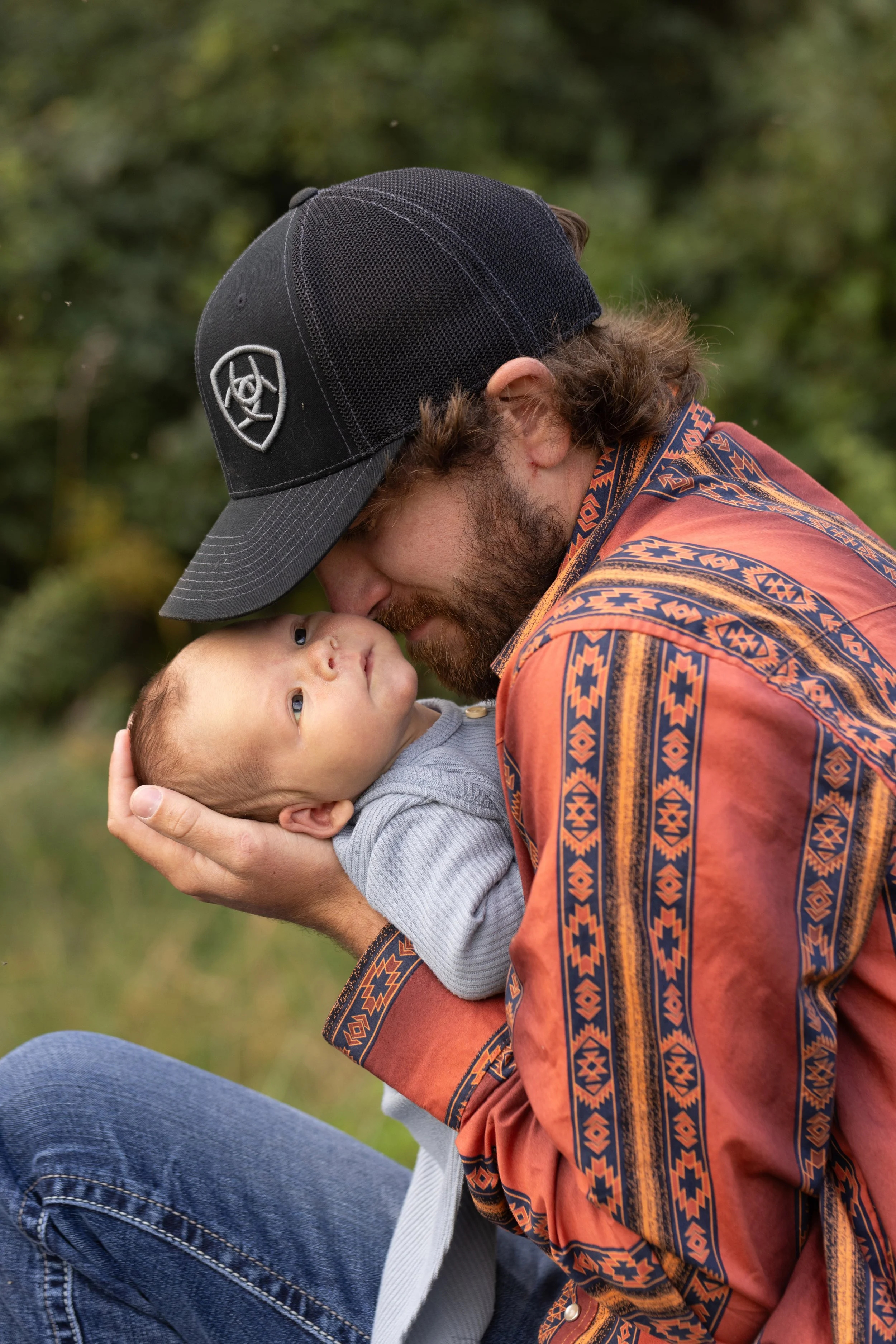 A man with a beard, wearing a patterned shirt and a black cap, holding a baby and leaning their foreheads together outdoors with green foliage in the background.