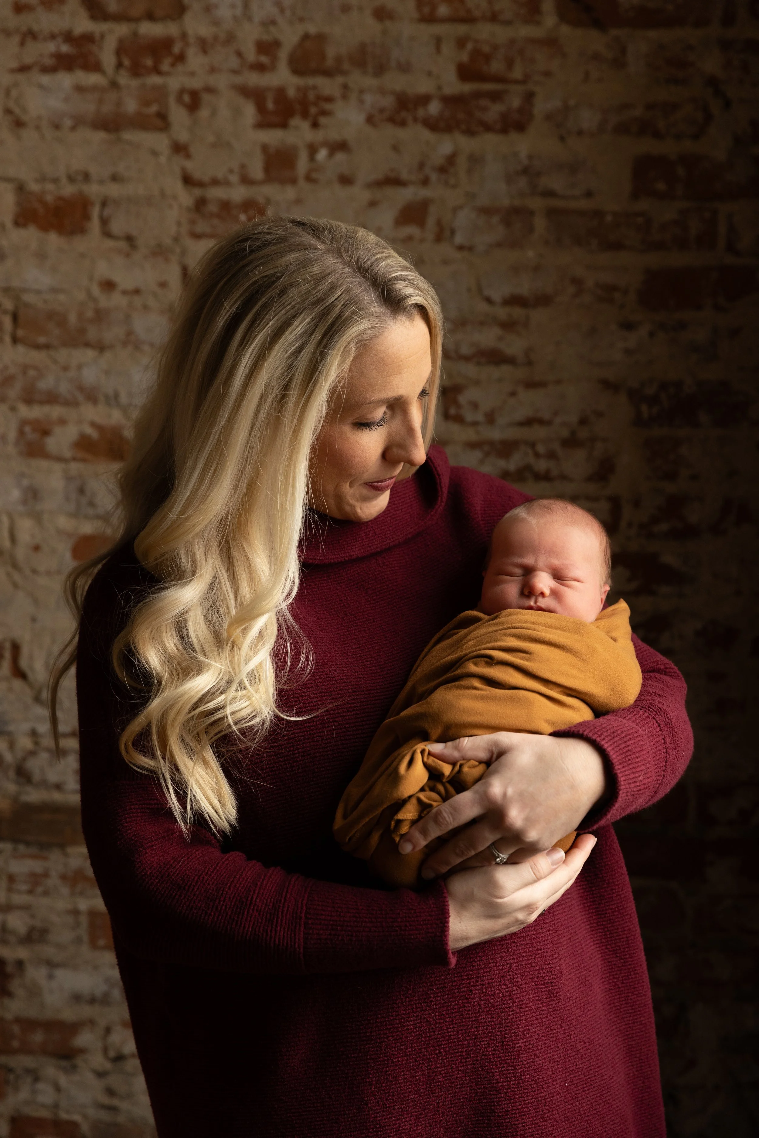A woman with long blonde hair holding a sleeping newborn wrapped in a mustard-colored blanket against a brick wall background.
