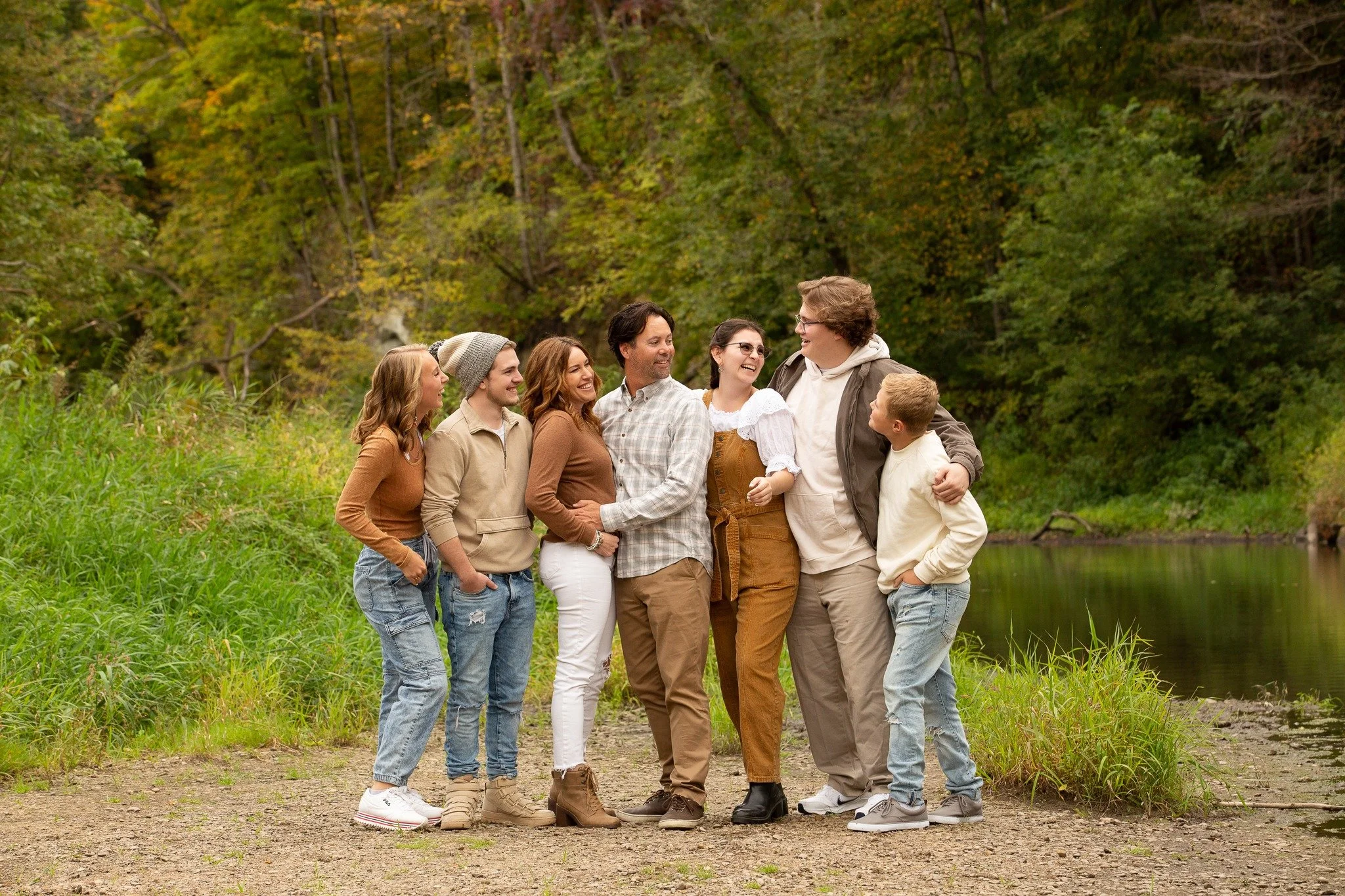 A family of eight people standing together outdoors near a river in a wooded area, smiling and looking at each other.