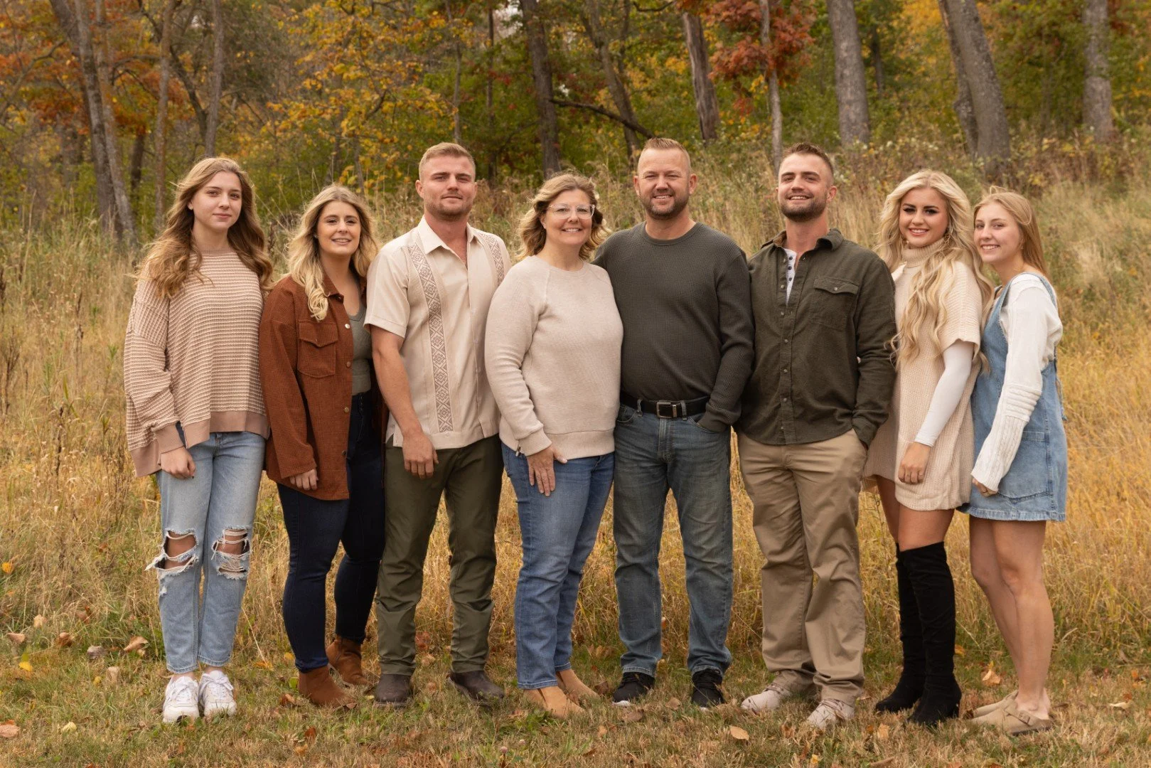 A family of nine standing outdoors in a field with autumn trees in the background, smiling at the camera.