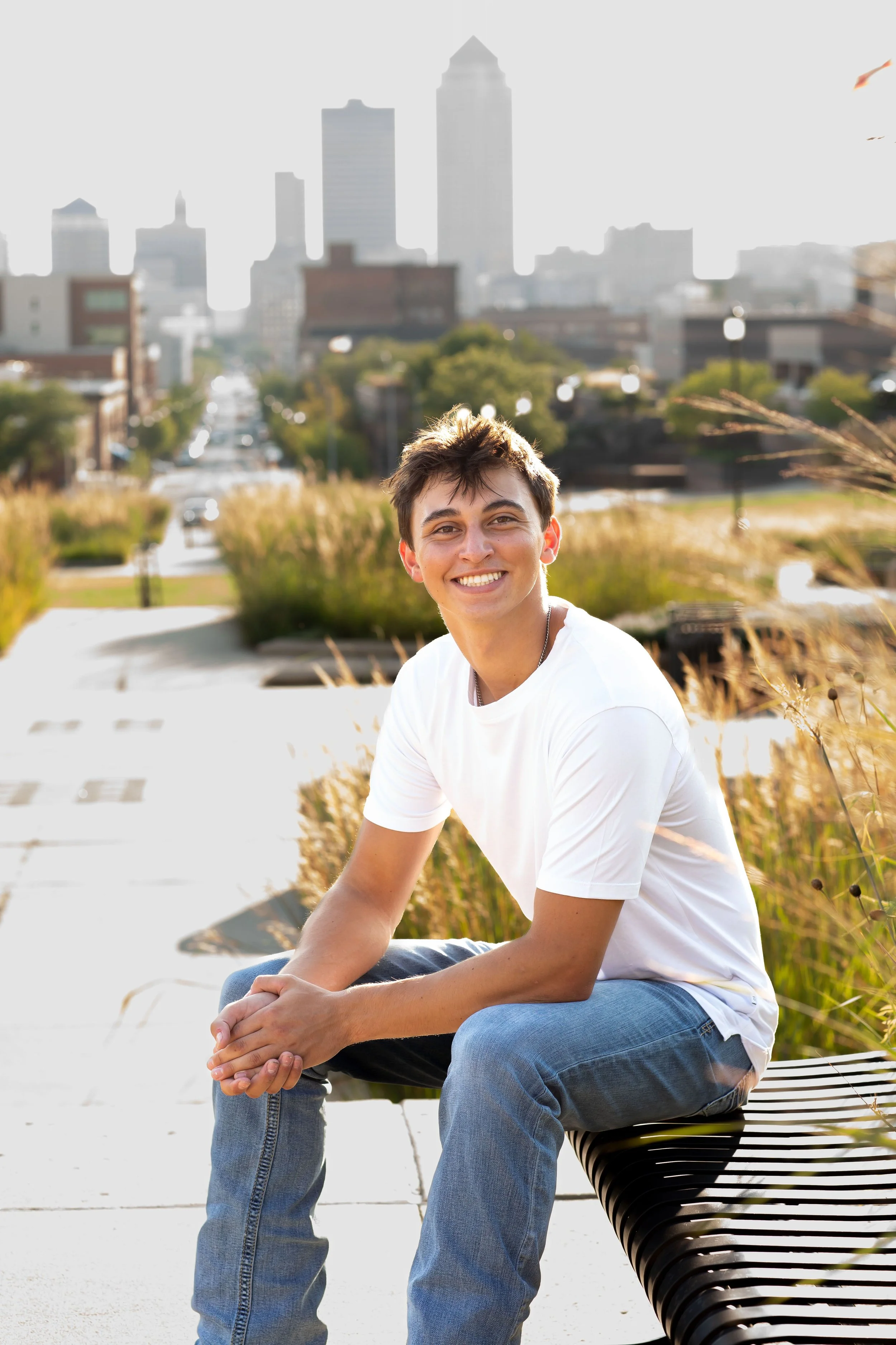A young man with short dark hair and a big smile, wearing a white t-shirt and blue jeans, sitting on a black bench outdoors with a city skyline in the background, during daytime.