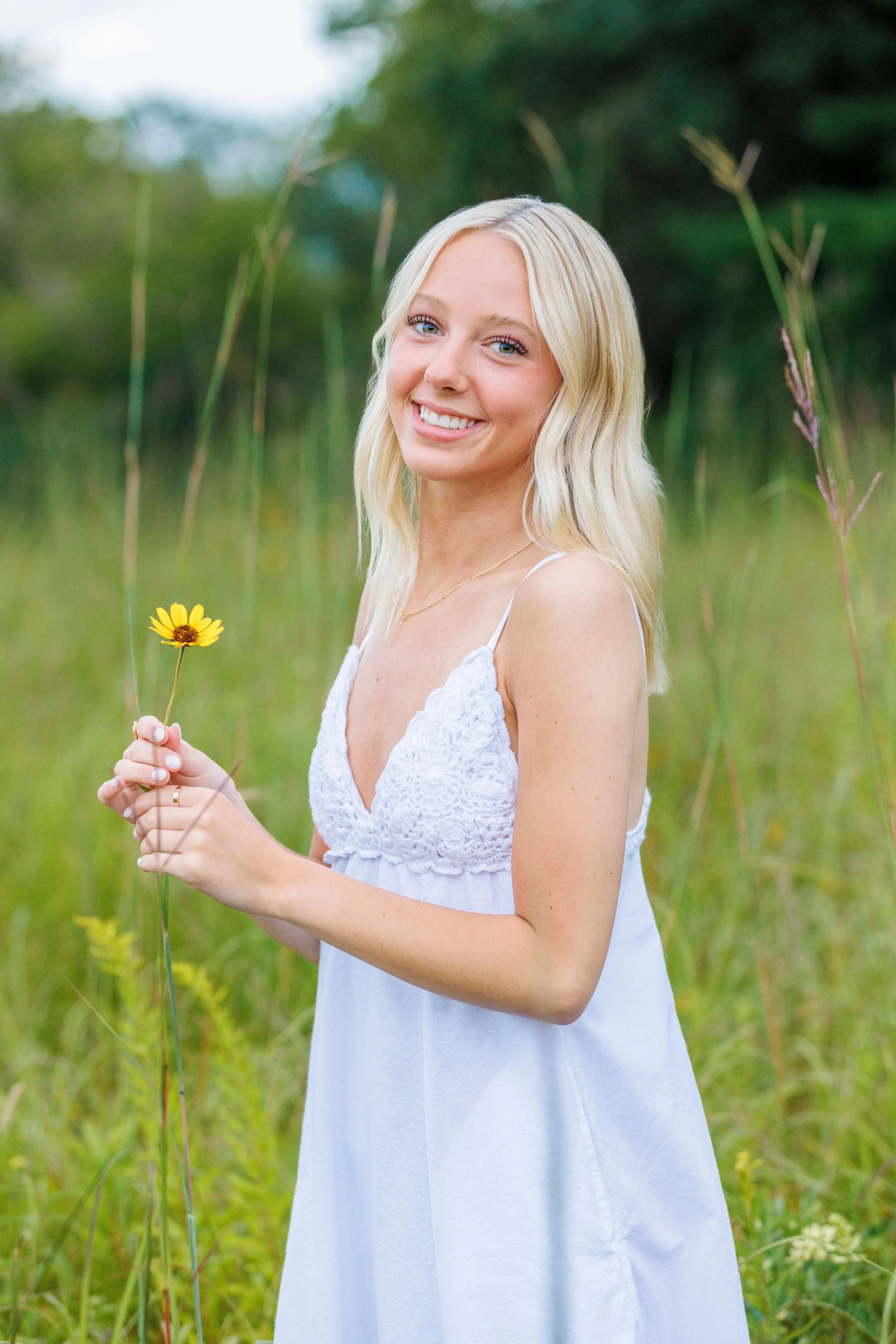 A young woman with blonde hair smiling and holding a yellow flower in a green field.