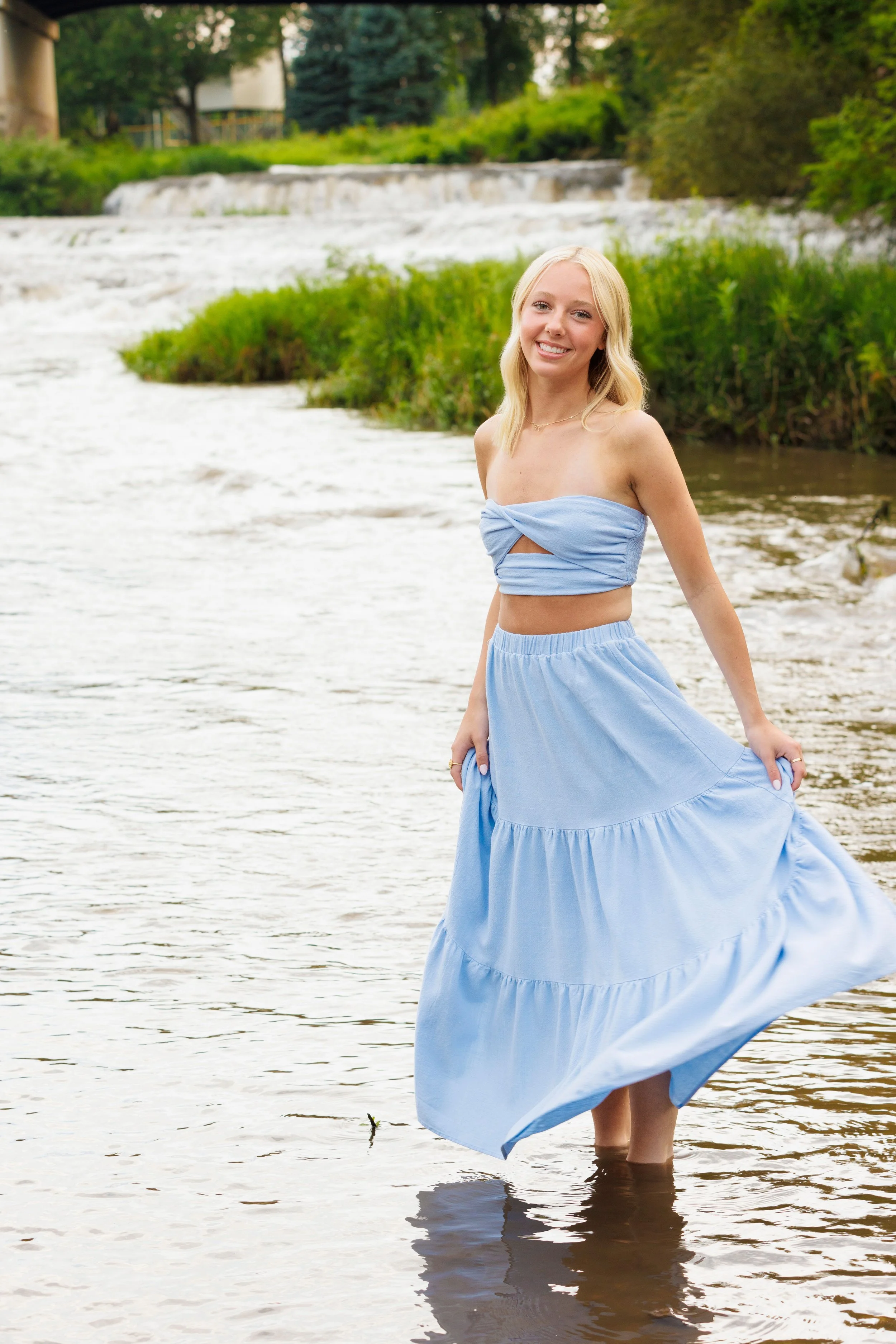 A young woman standing in a river with trees and a small waterfall in the background, smiling and wearing a light blue strapless dress.