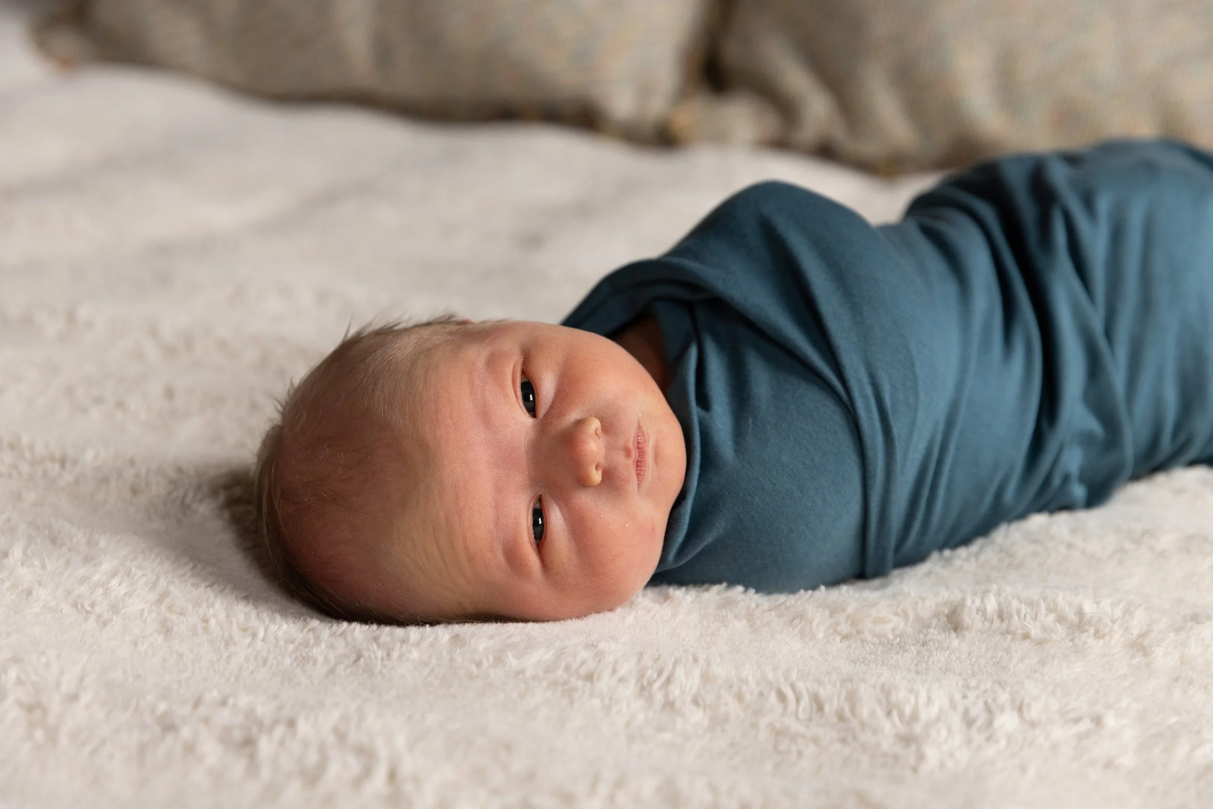 A newborn baby lying on a soft, light-colored blanket, wearing a teal shirt, looking at the camera with a calm expression.