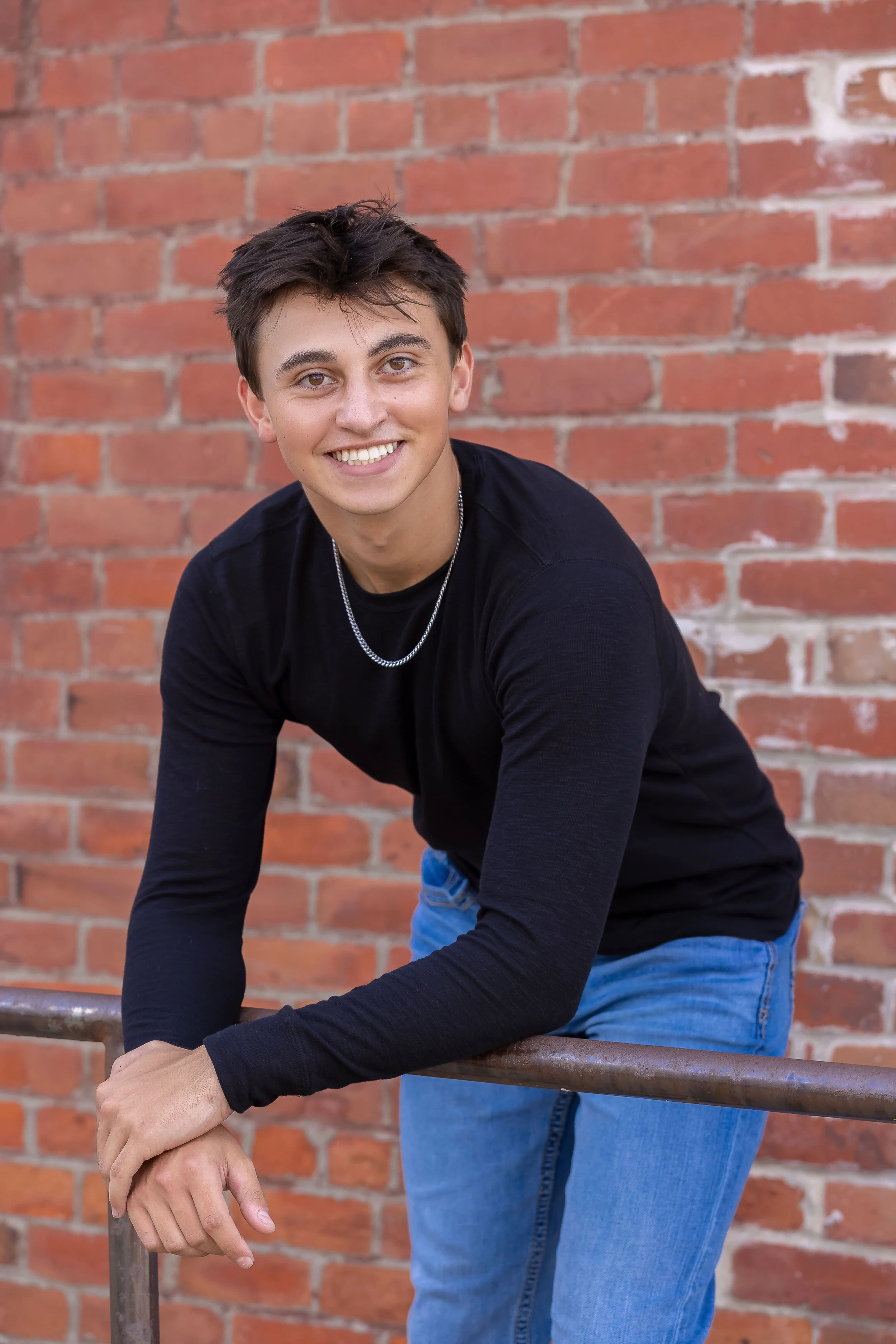 A young man with short dark hair, wearing a black long-sleeve shirt, blue jeans, and a silver chain necklace, smiling and leaning on a metal railing in front of a red brick wall.