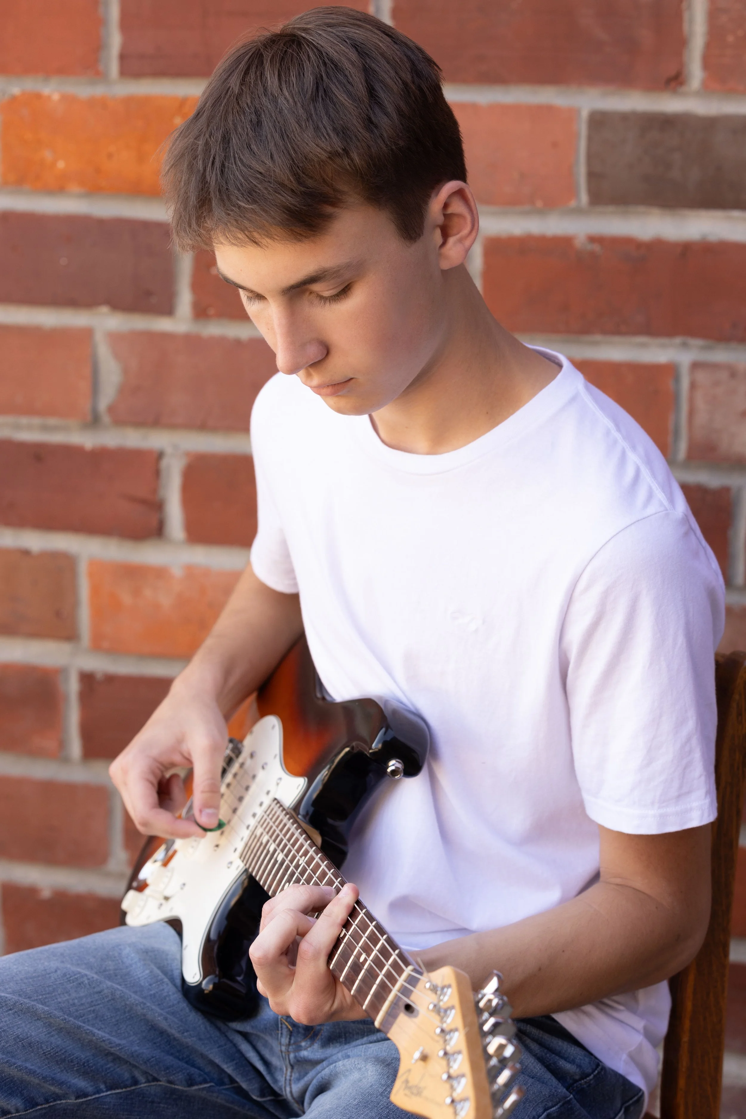 A young man in a white t-shirt and jeans sitting against a brick wall while playing an electric guitar.