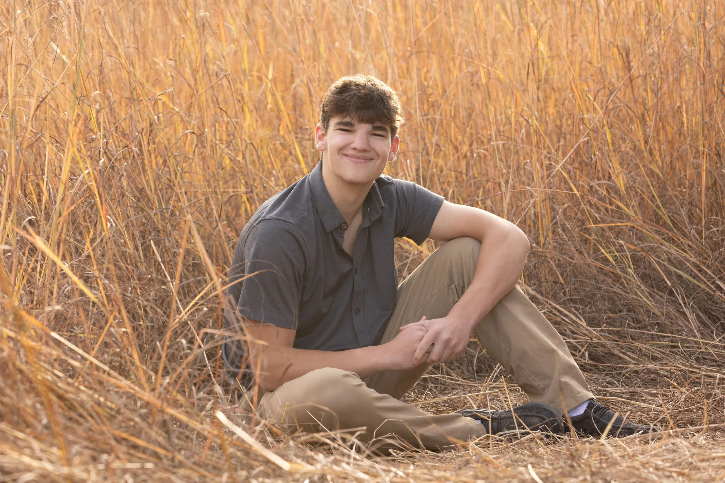 A young man sitting on the ground in a field of tall grass, smiling at the camera during sunset.