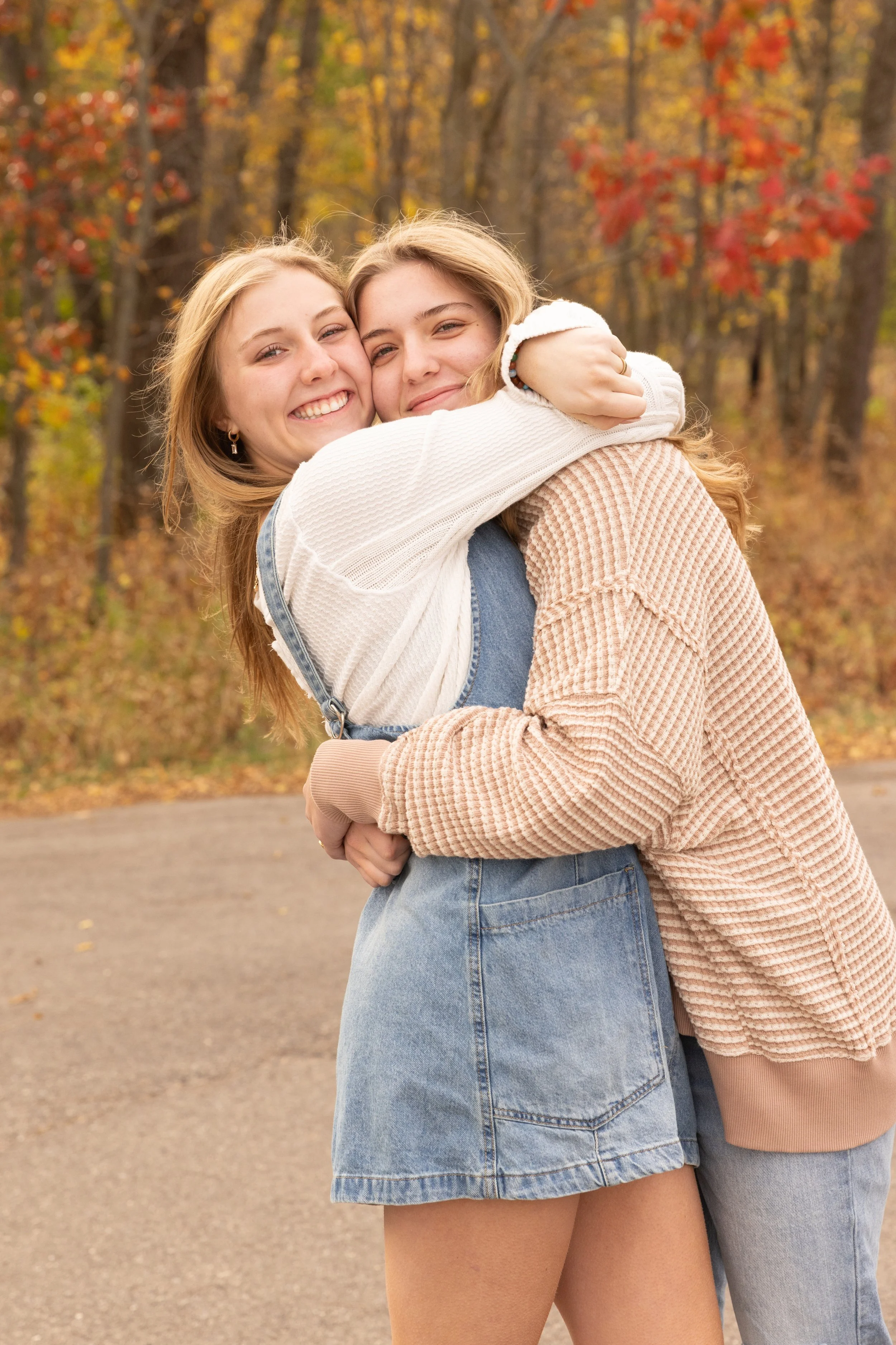 Two young women hugging outdoors during autumn, with trees showing fall foliage in the background.