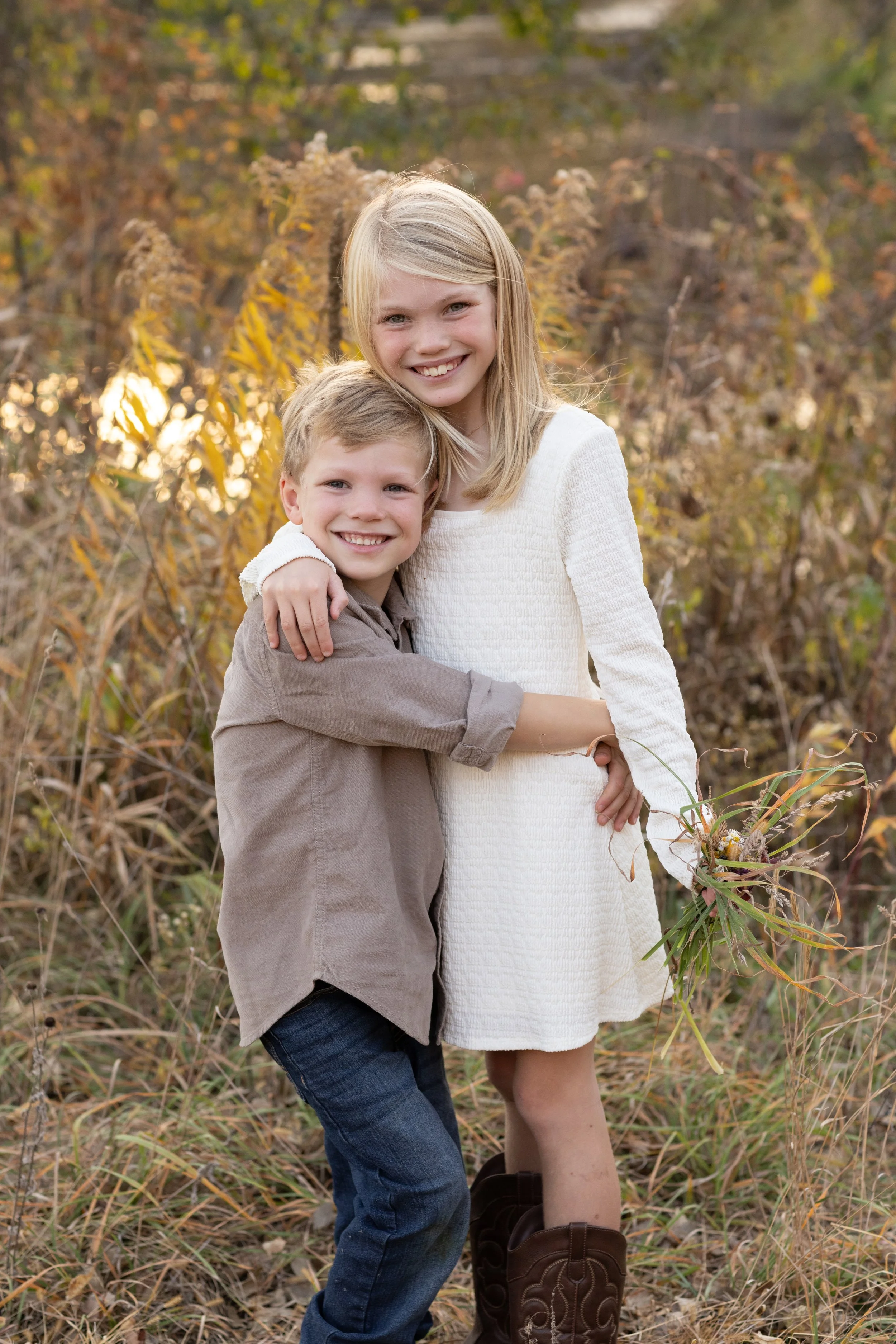 Smiling young girl in a white dress and a young boy in a gray shirt hugging outdoors during fall with yellow and brown foliage in the background.