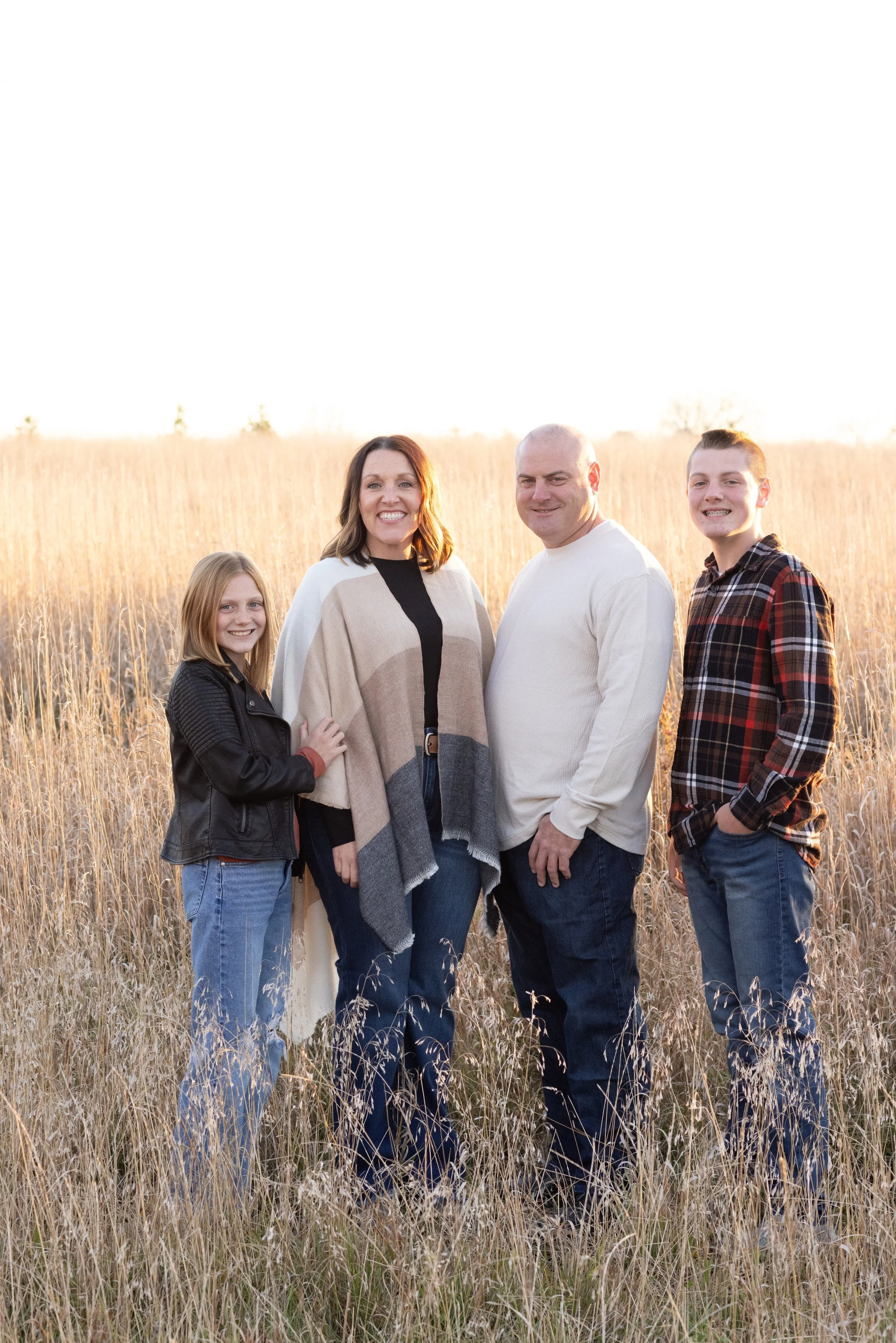 A family of four standing in a field of tall, dry grass during sunset, smiling at the camera.