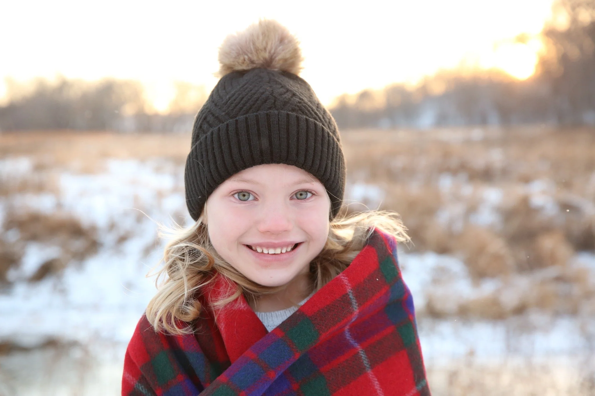 A young girl with blonde hair, blue eyes, and a big smile, wrapped in a red and green plaid blanket, wearing a black knit beanie with a pom-pom on top, standing outdoors in a snowy field during sunset.