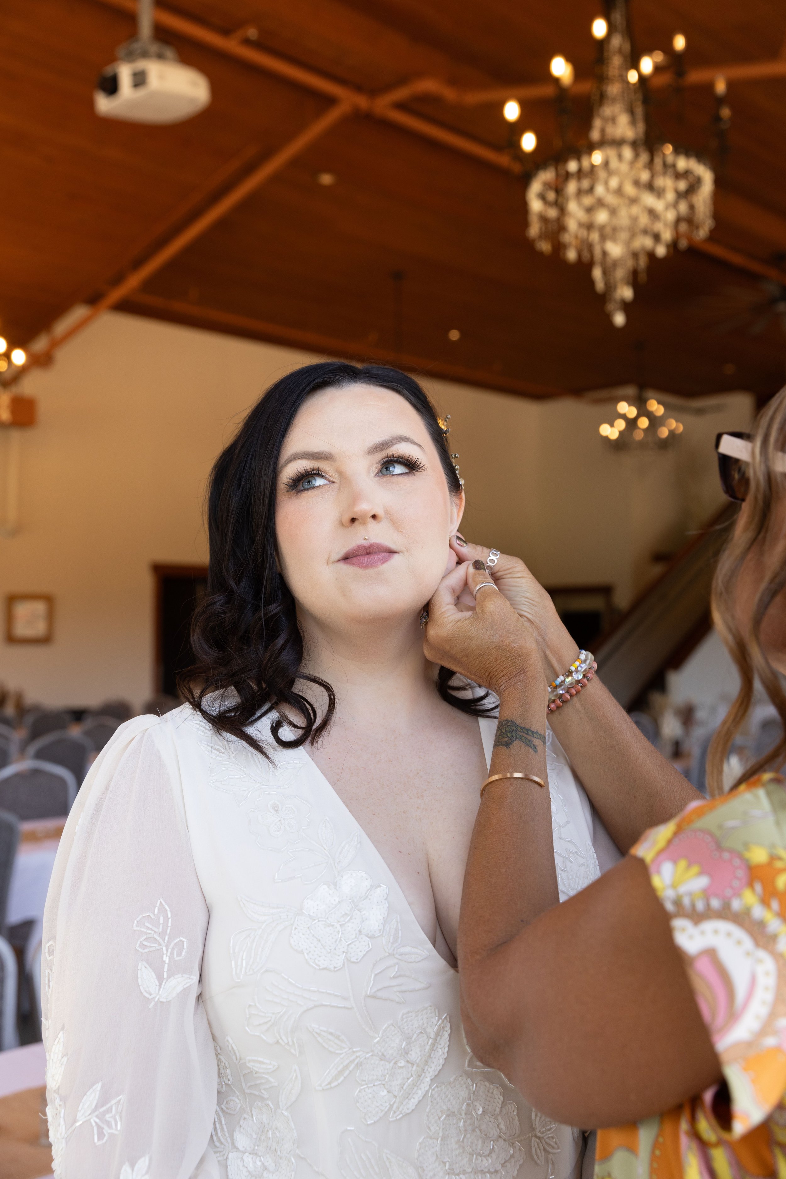 A woman with black hair and blue eyes, wearing a white embroidered dress, getting her earrings adjusted by another woman with a tan skin tone and colored bracelets, inside a decorated event hall with chandeliers and a wooden ceiling.
