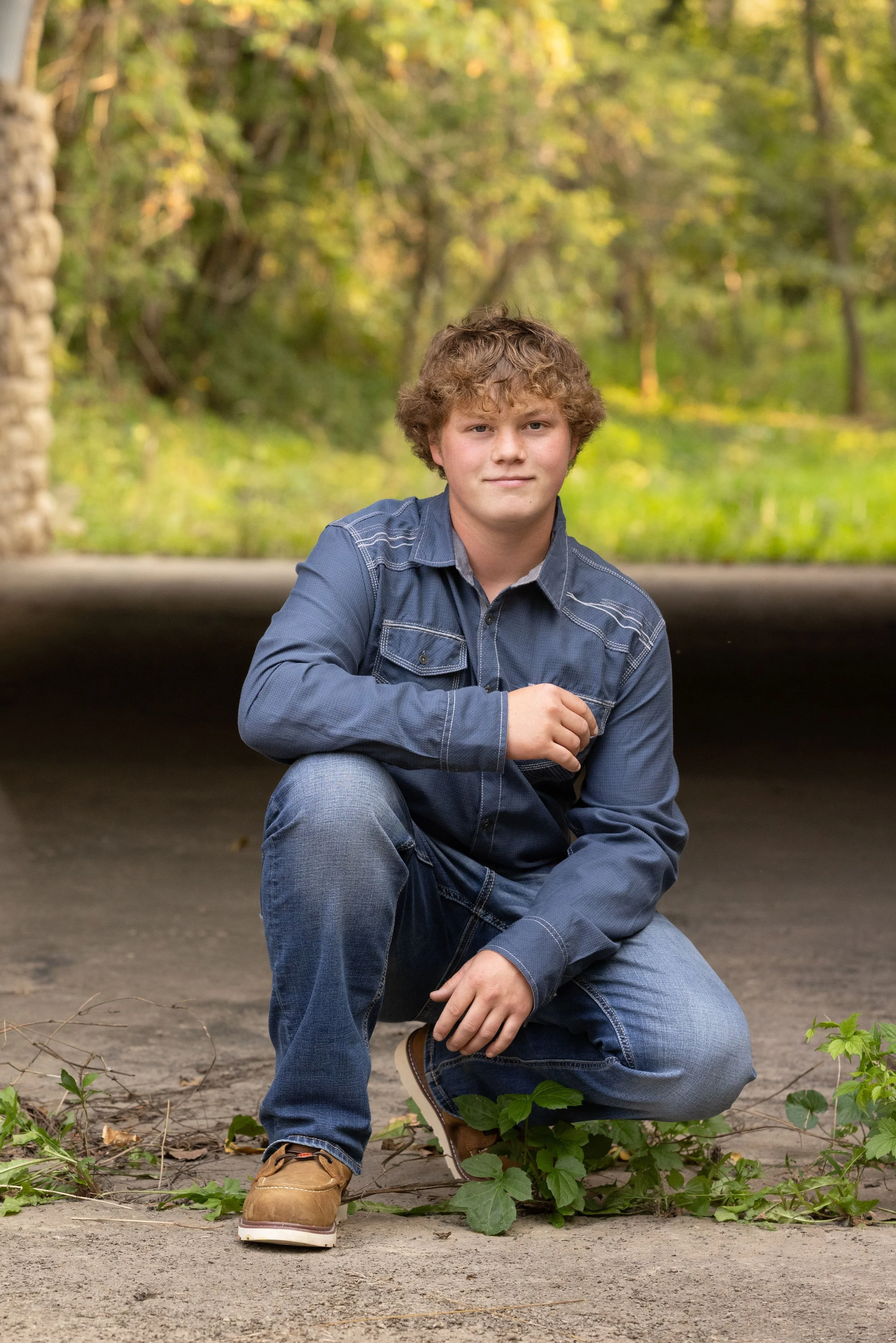 A young man with curly hair, wearing a denim shirt and jeans, kneeling on one knee outdoors on a paved surface with greenery and trees in the background.