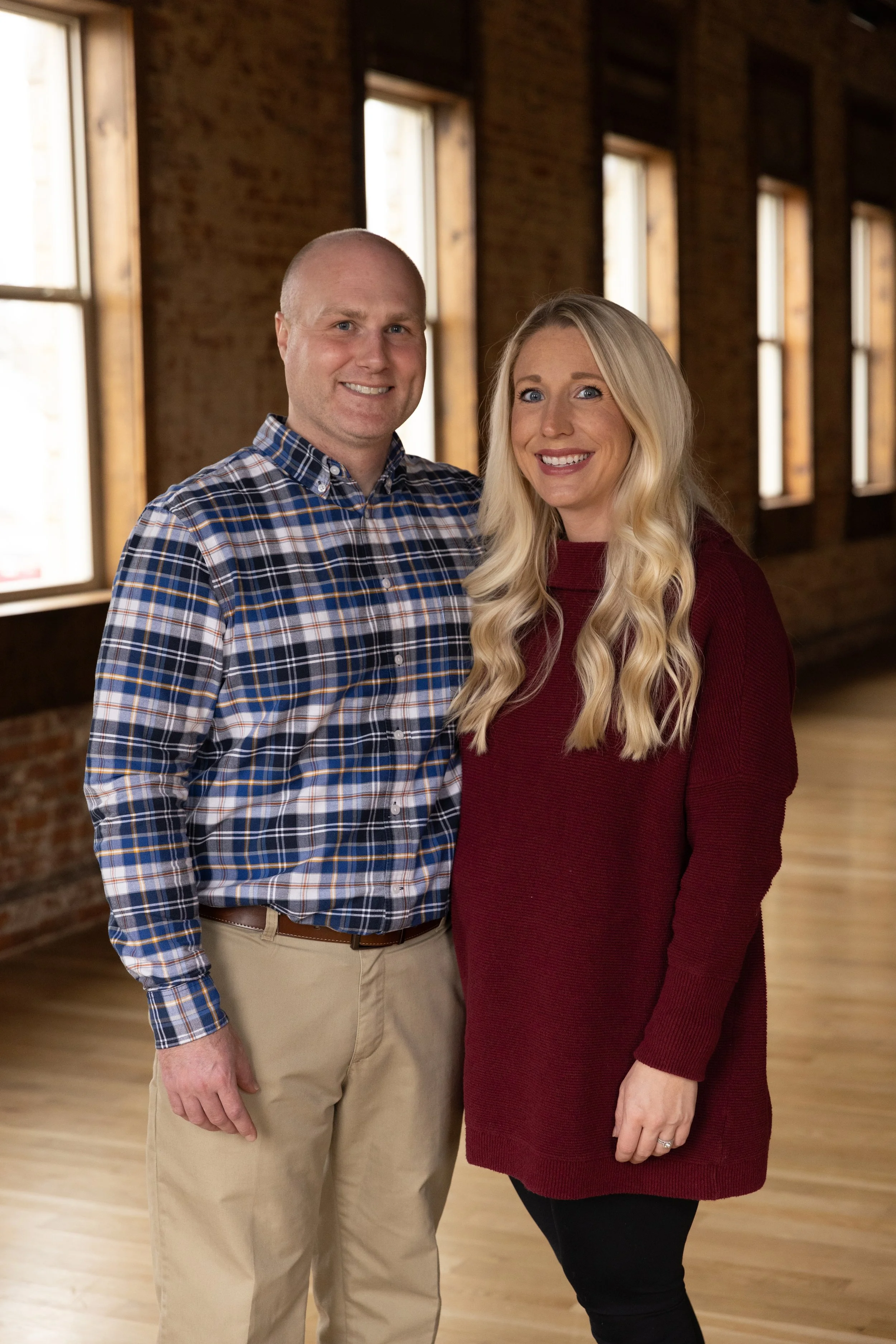 A man and woman standing indoors near windows, smiling at the camera.