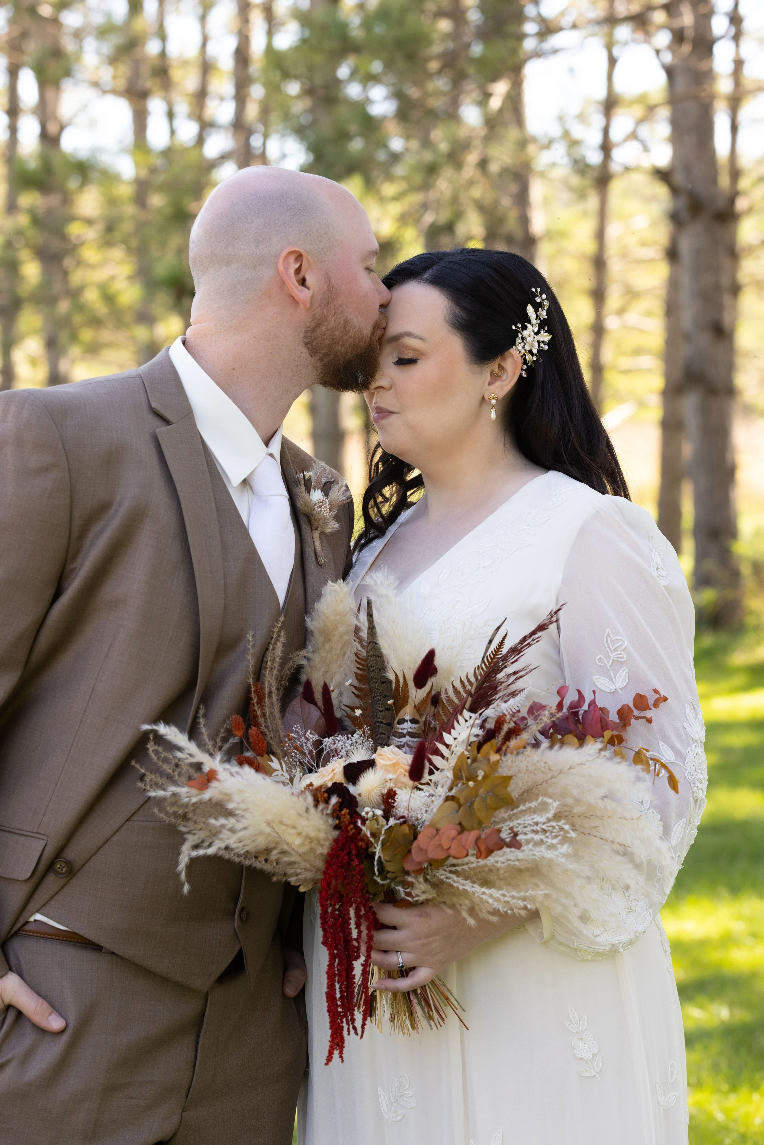 A bride and groom sharing a tender moment outdoors, with the groom kissing the bride's forehead, holding a bouquet of dried flowers.