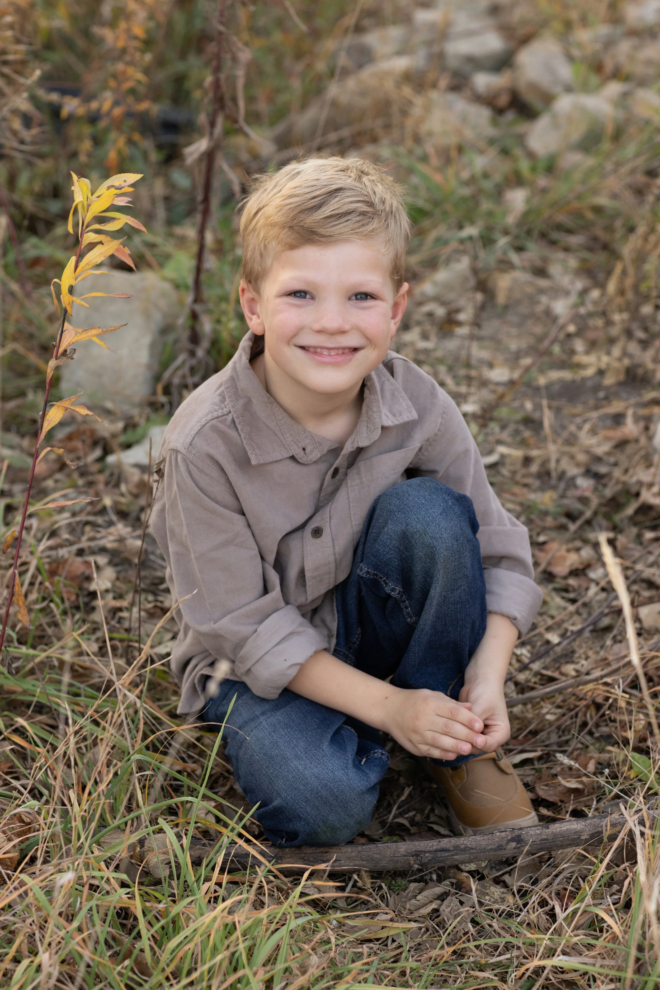 A young boy with blonde hair, blue eyes, and a light brown shirt sitting on the ground outdoors among fall leaves and grass, smiling at the camera.