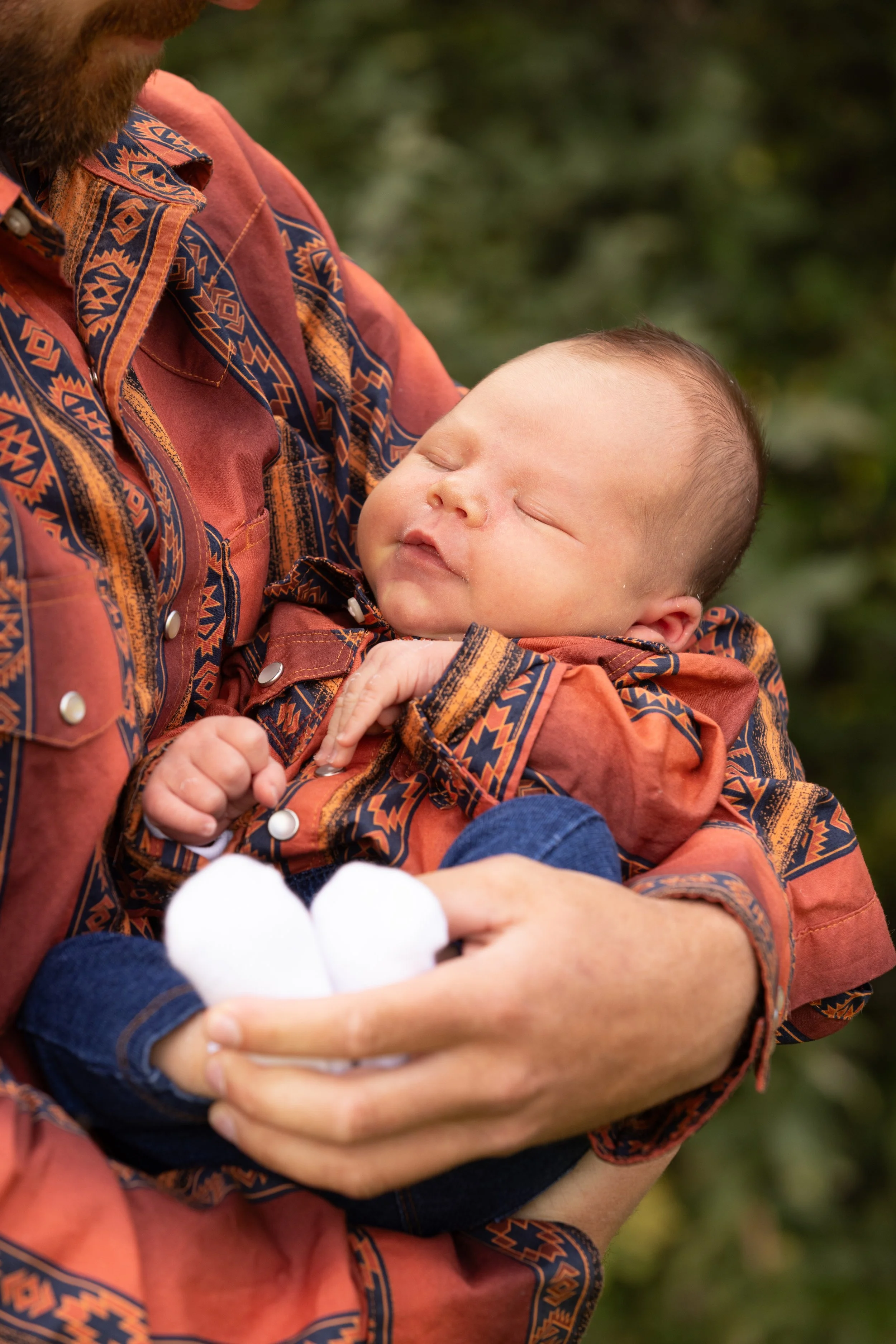A person holding a sleeping baby in their arms, with the baby resting their head on the person's chest. The baby has its eyes closed, and the person is wearing a patterned, reddish-brown shirt.