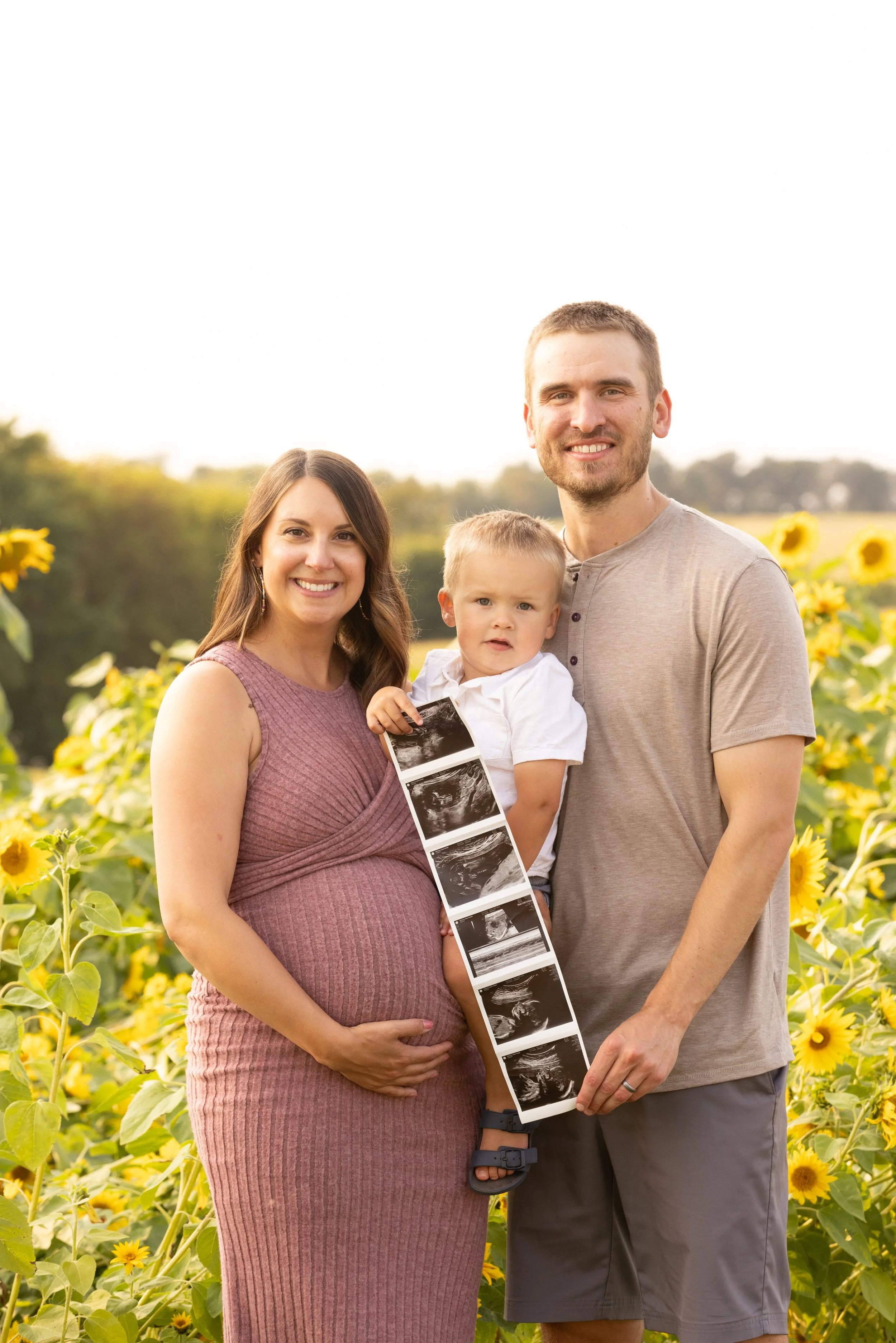 A pregnant woman, a man, and a young boy holding ultrasound images stand in a sunflower field during daytime, smiling at the camera.