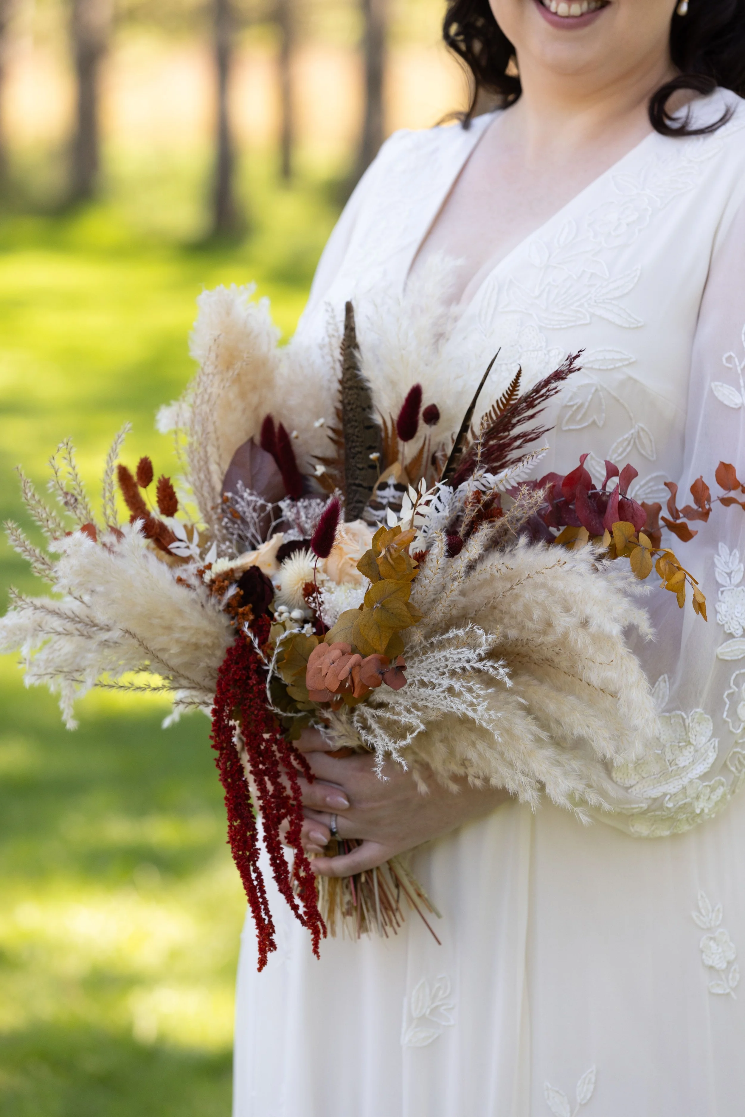 A woman in a white dress holding a bouquet of dried flowers including pampas grass, preserved leaves, and other botanicals, outdoors in sunlight.