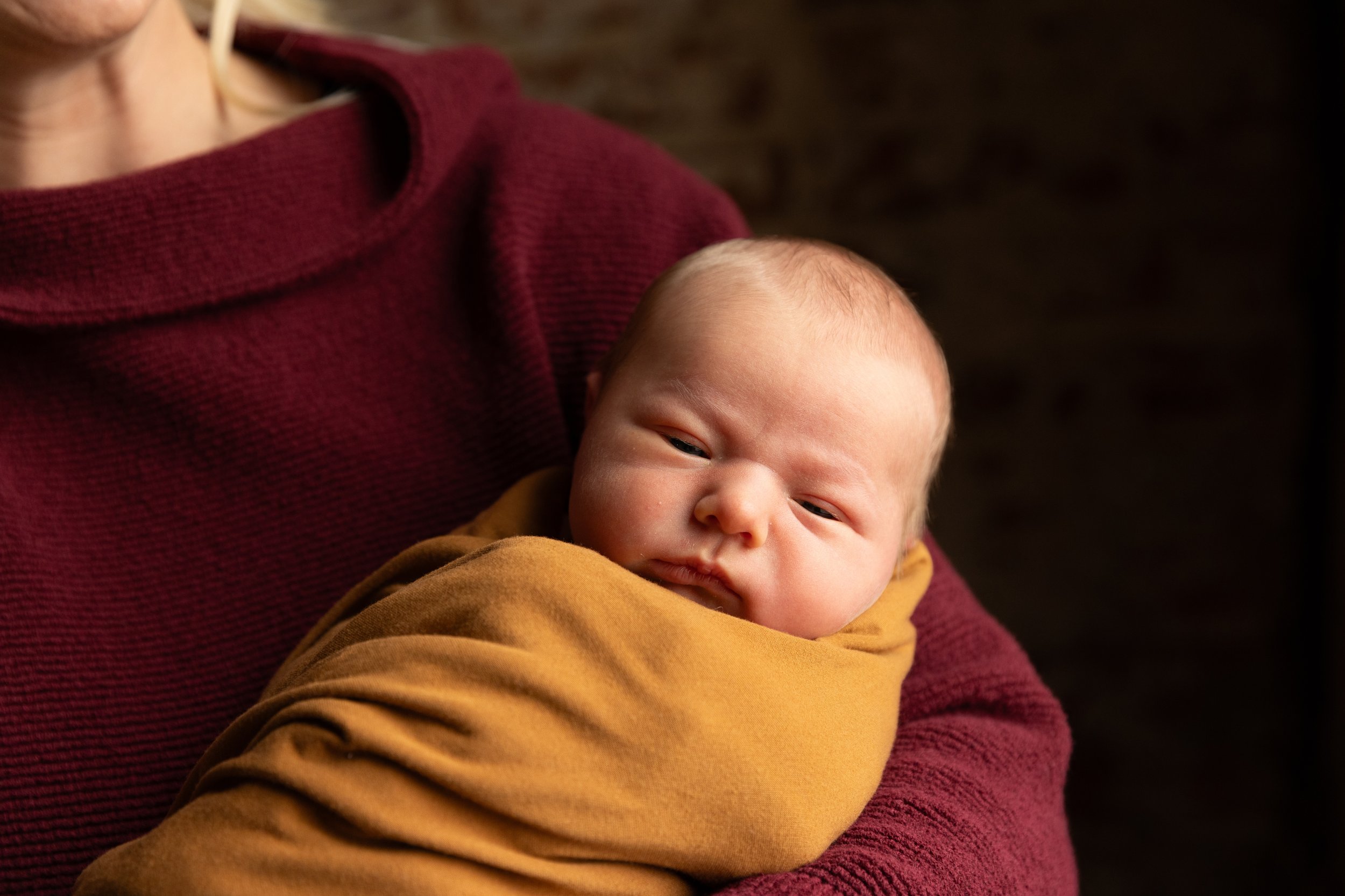 A person holding a young baby wrapped in a mustard-colored blanket, with a dark background.