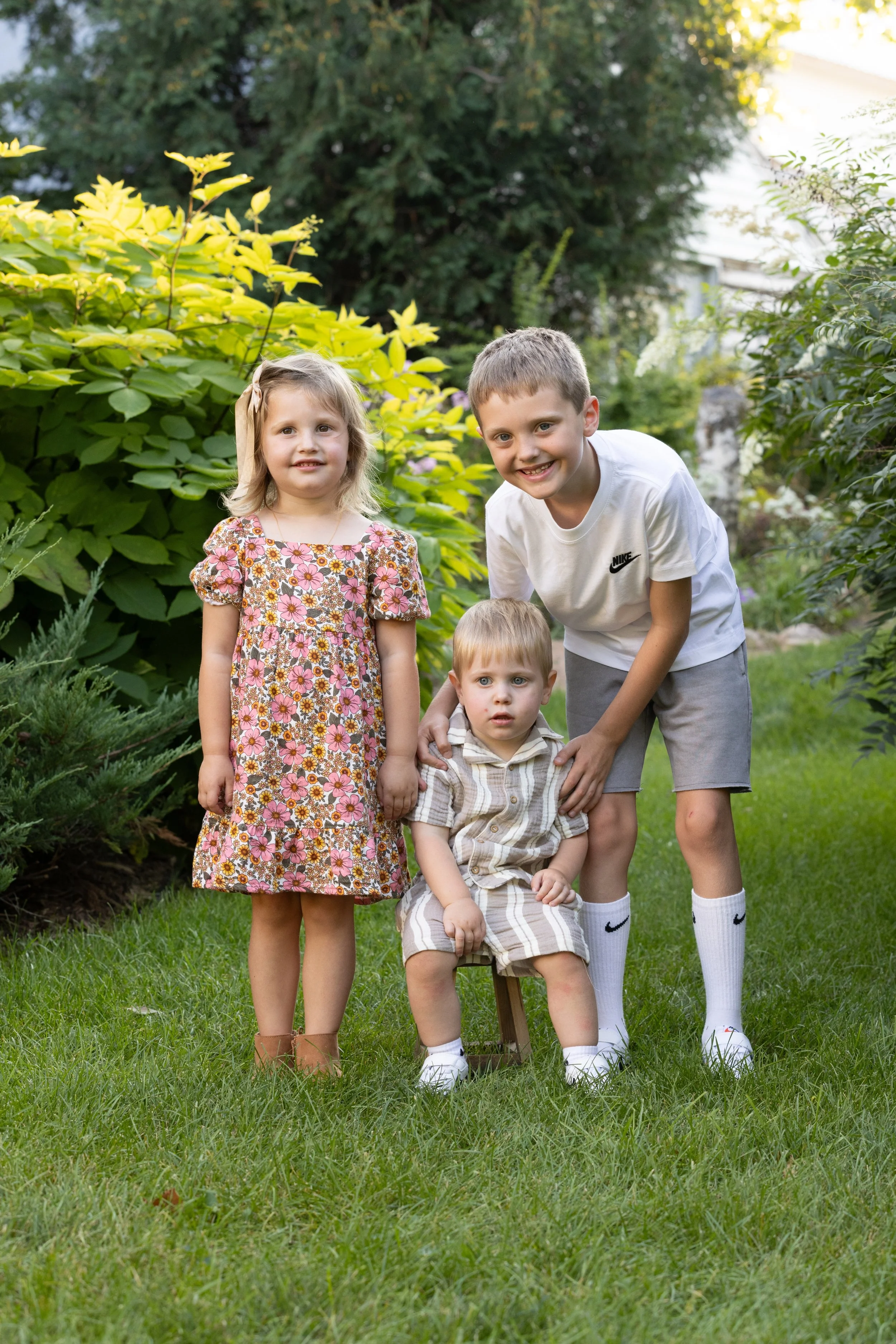 Three young children, two boys and a girl, posing outdoors in a garden with green foliage. The girl is standing on the left wearing a floral dress, the older boy is crouching on the right wearing a white Nike T-shirt, and the younger boy is sitting o