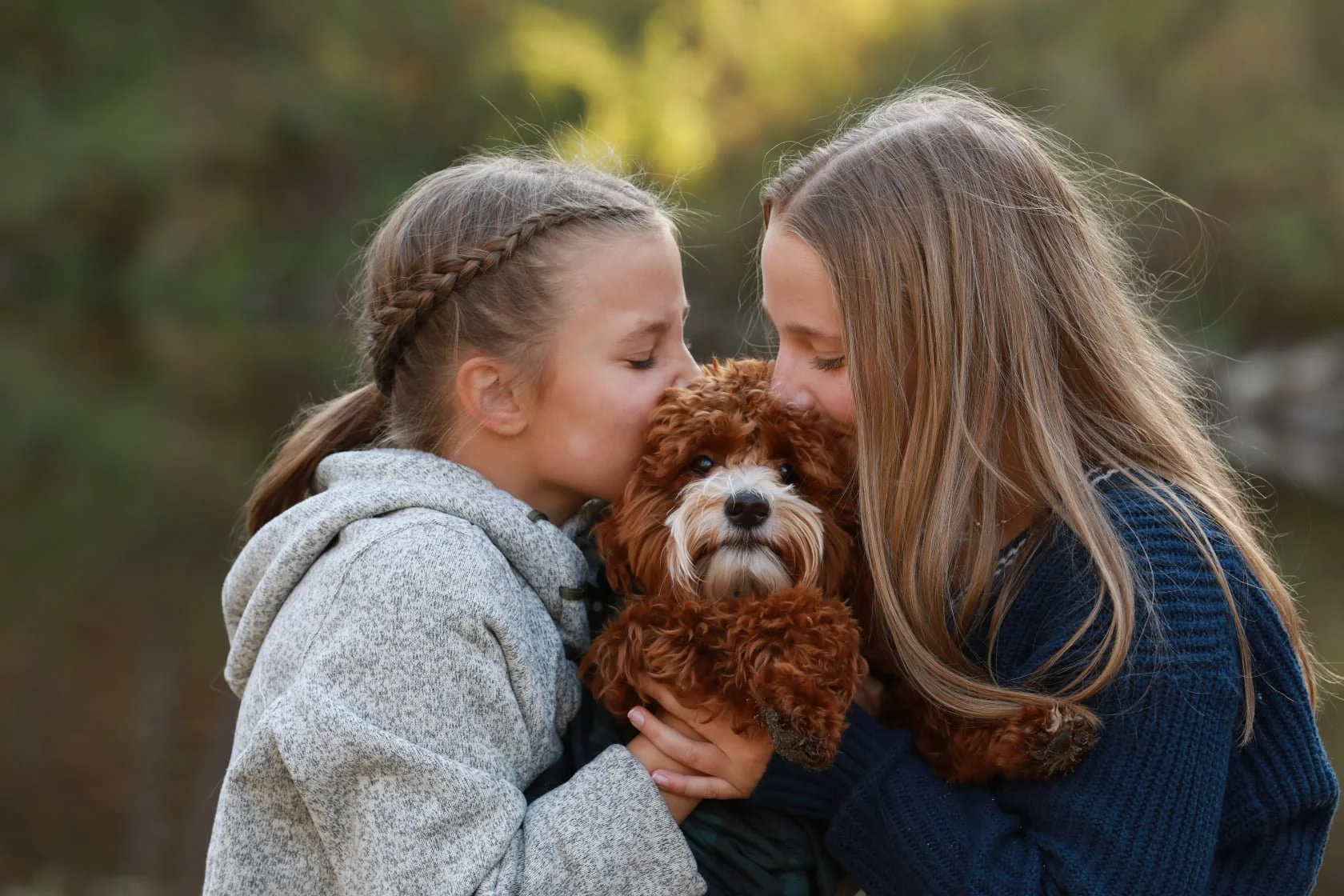 Two girls kissing a toy poodle puppy outdoors.