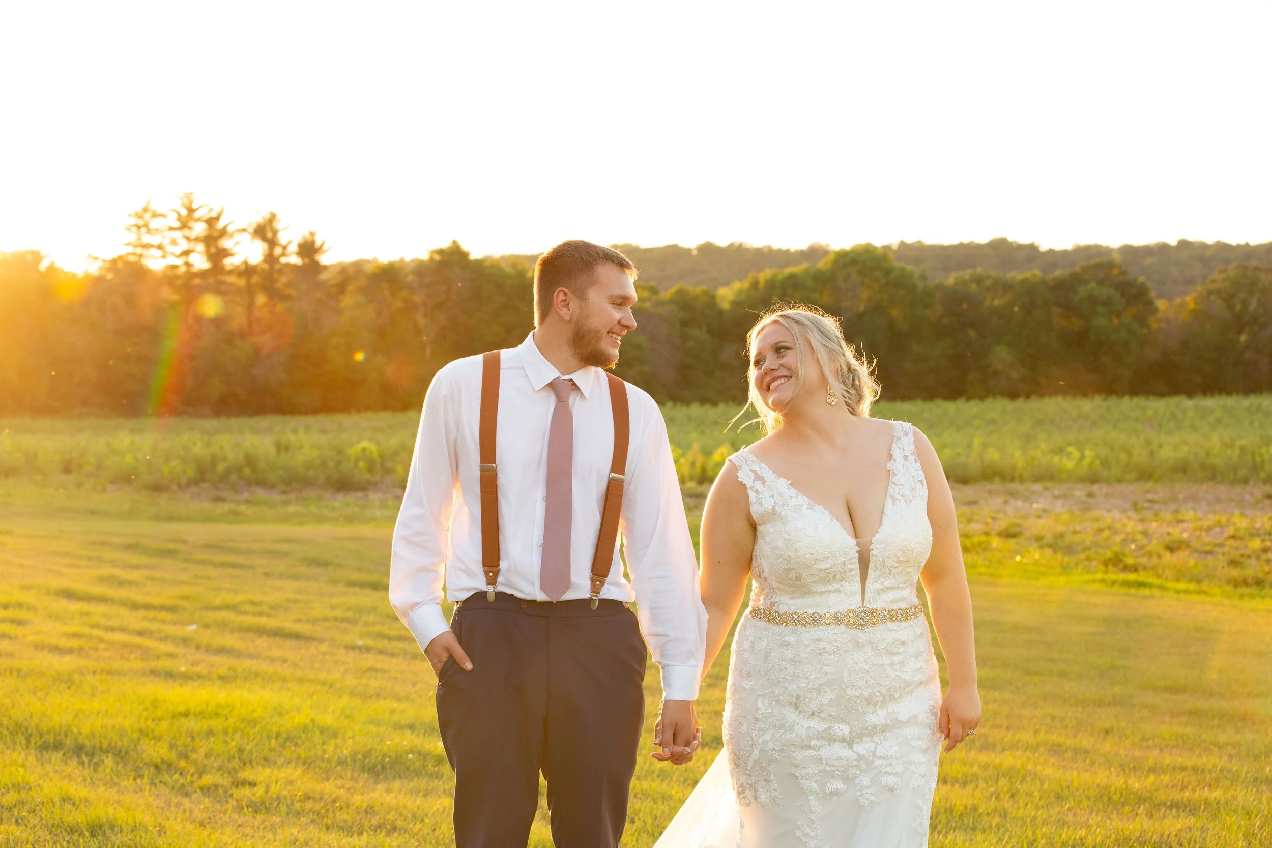 A newlywed couple walking hand-in-hand outdoors during sunset, with a green field and trees in the background.