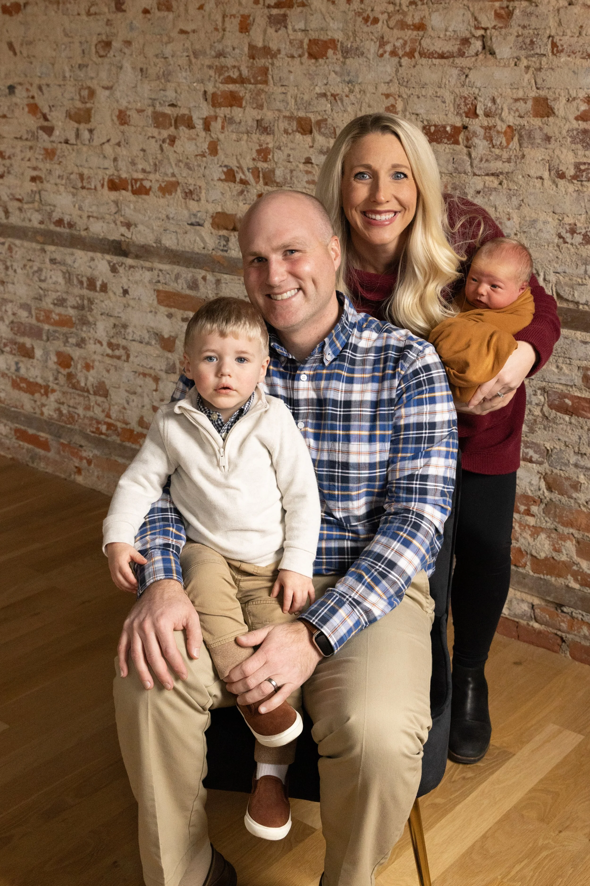 A family of four posing in front of an exposed brick wall, including a man, a woman holding a newborn, and two young boys.