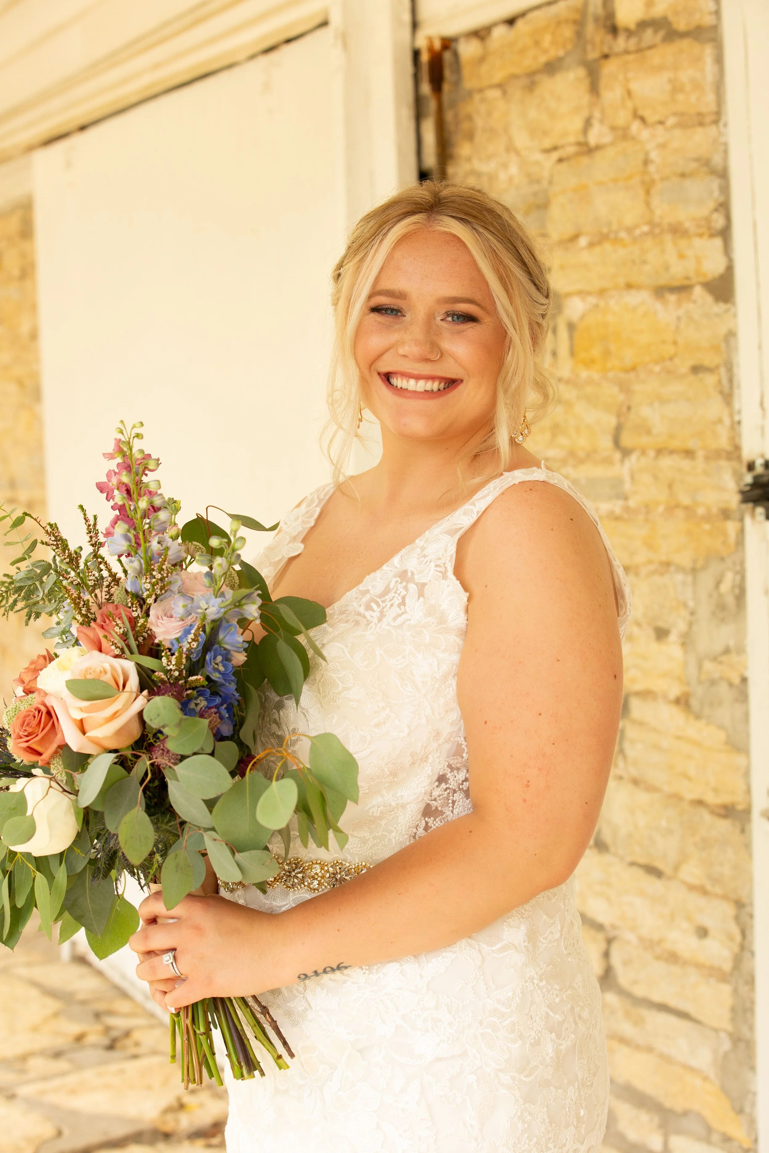 A woman in a wedding dress holding a bouquet of flowers, smiling at the camera, standing against a rustic stone wall background.