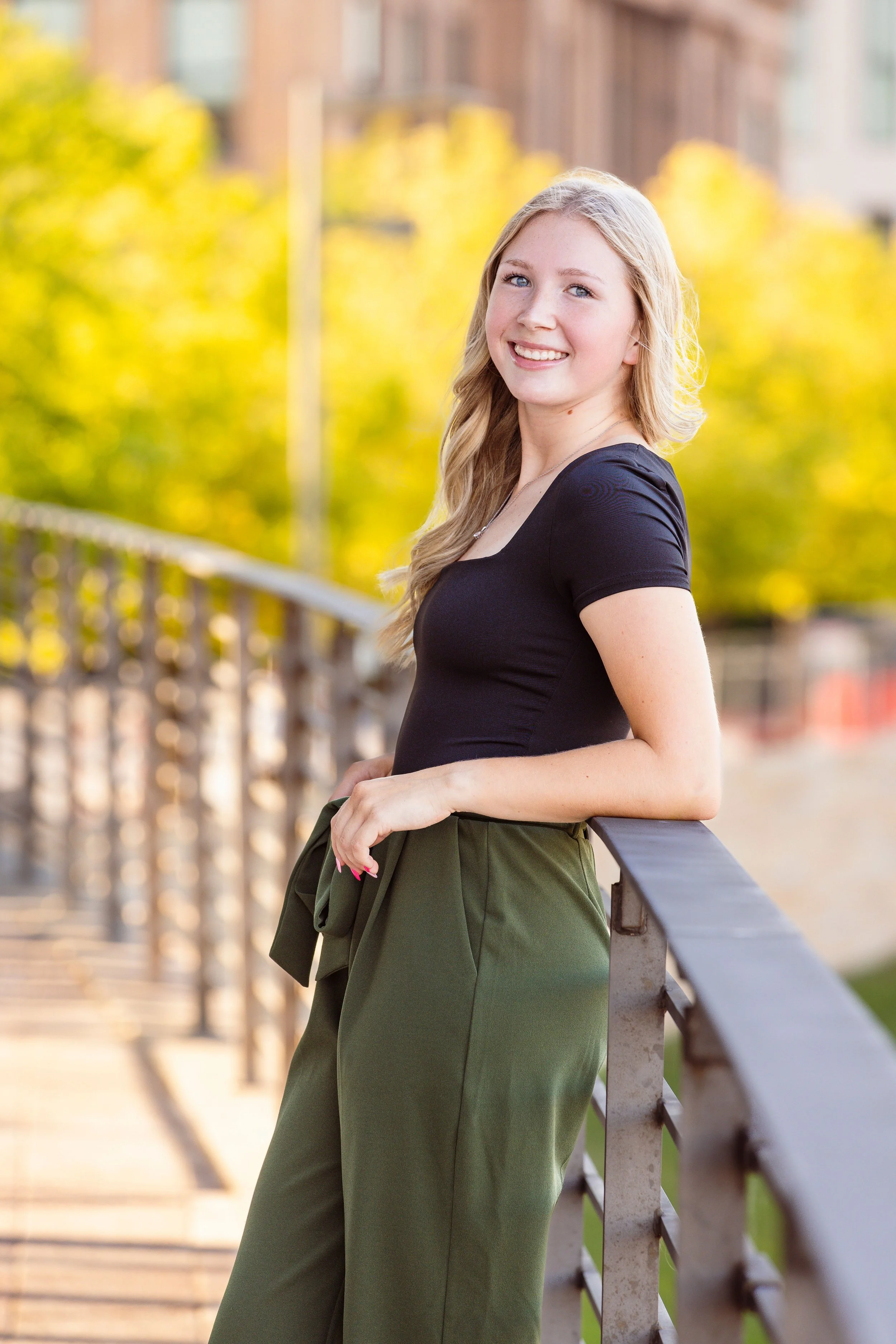 A young woman with long blonde hair and blue eyes smiling and leaning on a railing outdoors with trees and buildings in the background.