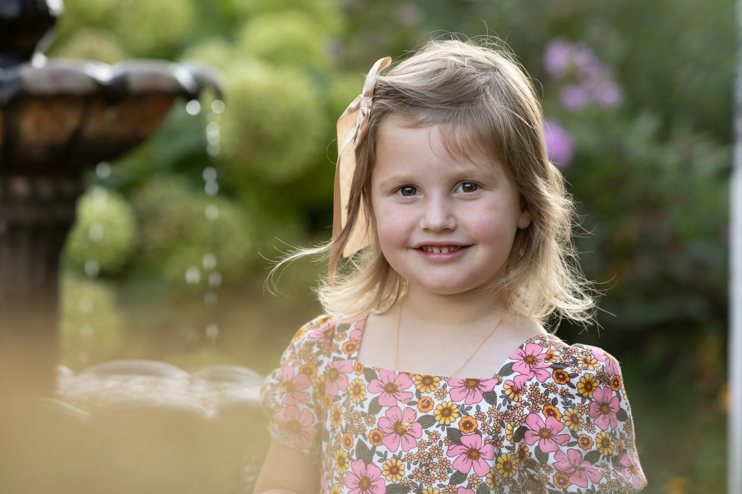 A young girl with blonde hair, wearing a pink floral dress and a ribbon in her hair, smiling outdoors with a blurred garden background.