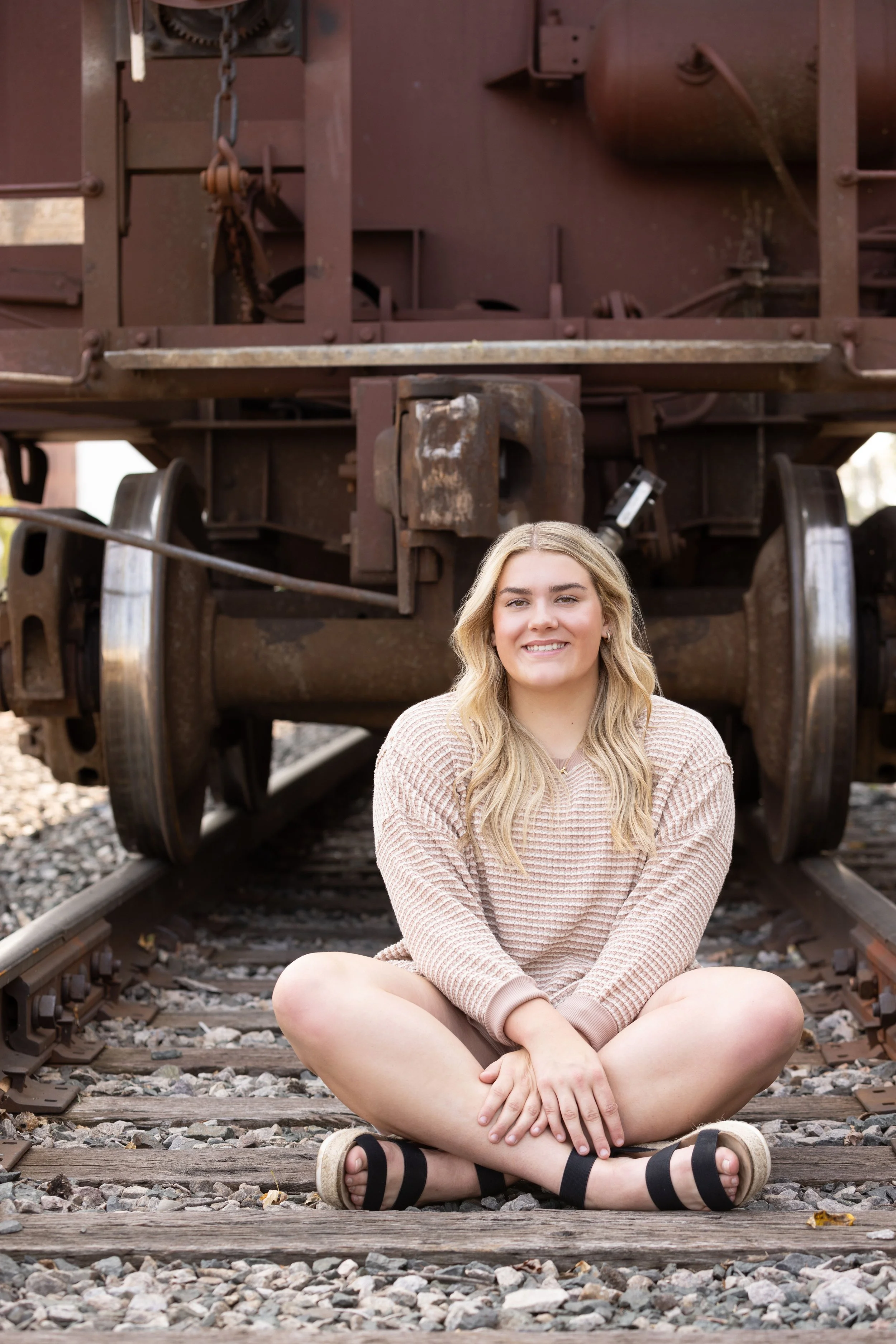 A young woman with long blond hair sitting cross-legged on railroad tracks in front of an old rusted train car, smiling at the camera.
