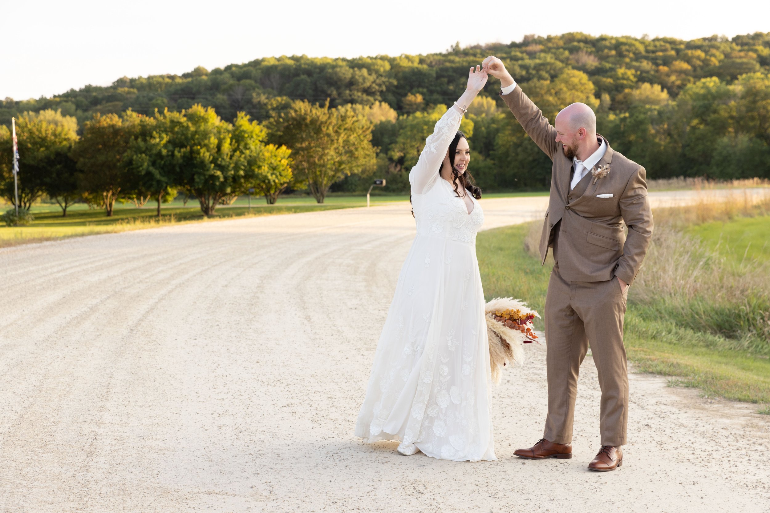 A bride and groom dancing outdoors on a dirt road with trees and hills in the background during sunset.
