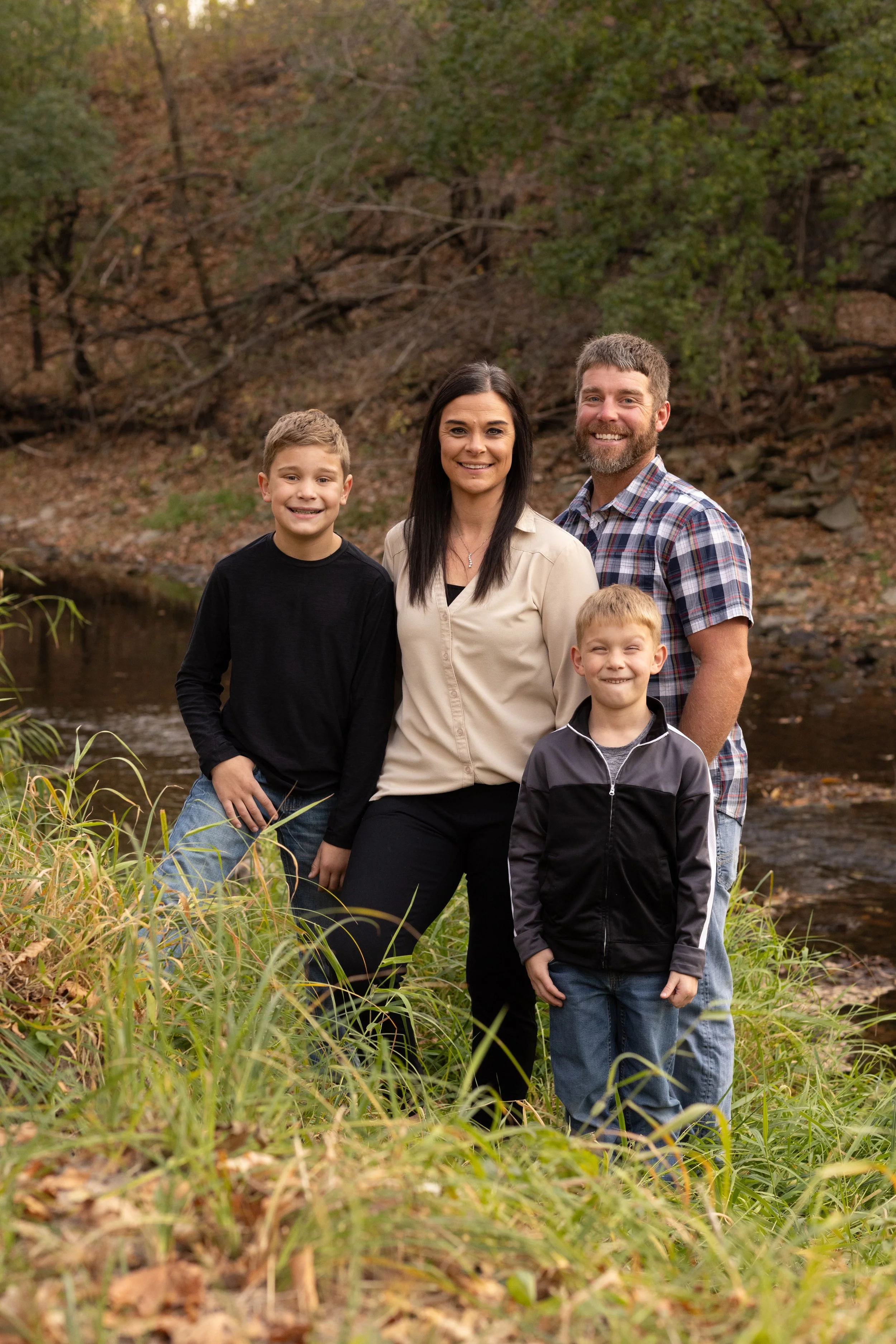 A family of five stands outdoors near a creek with trees and rocks in the background, smiling at the camera.