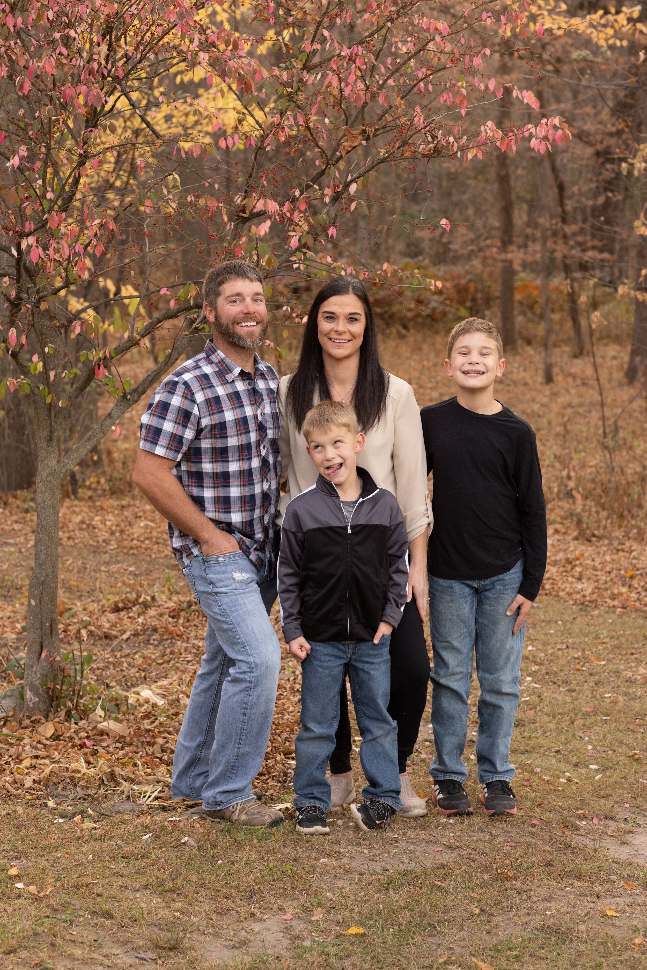 A family of four standing outdoors in fall, next to a pink-leafed tree, smiling at the camera.