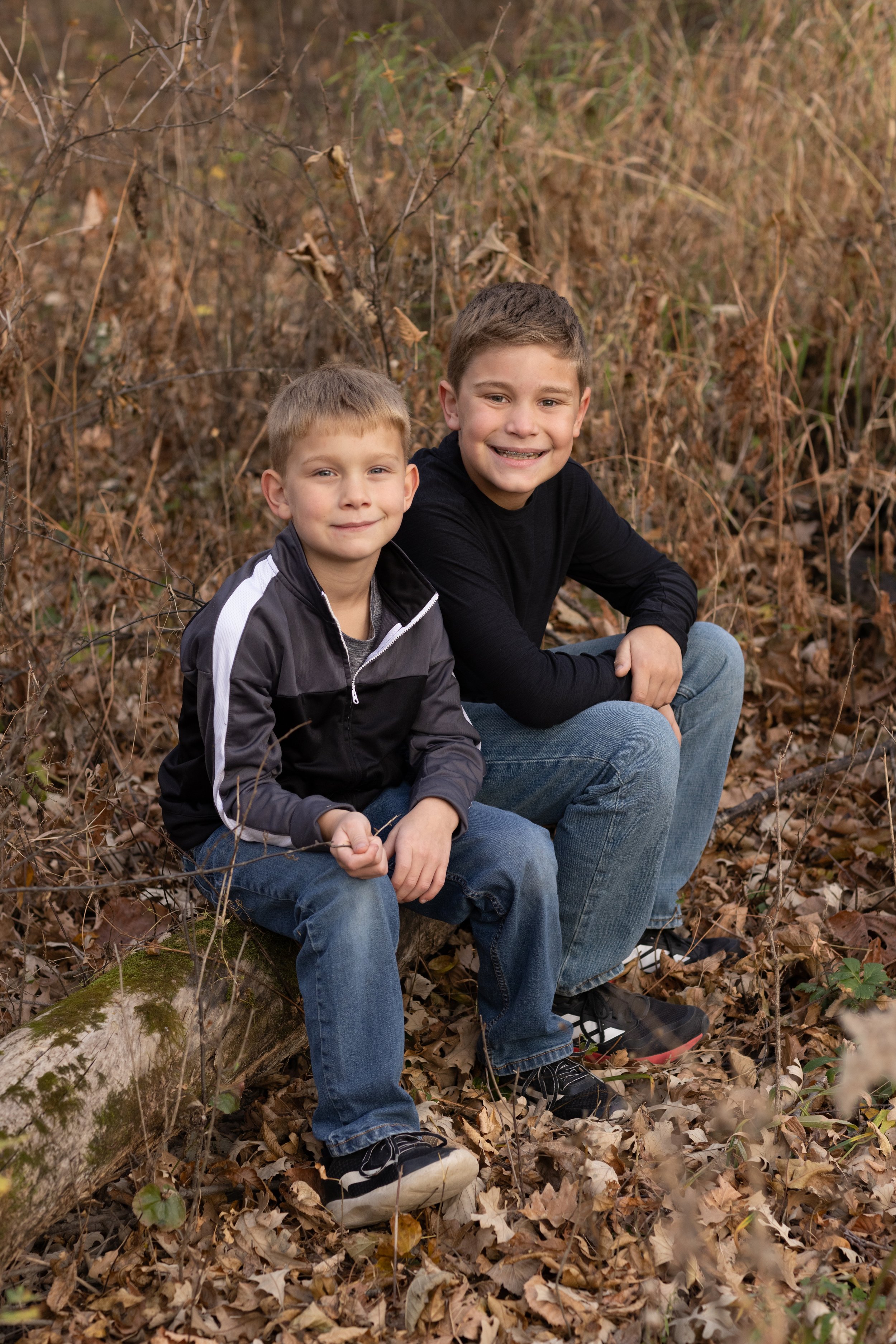 Two boys sitting on a fallen tree in a forest with dry leaves on the ground, smiling at the camera.