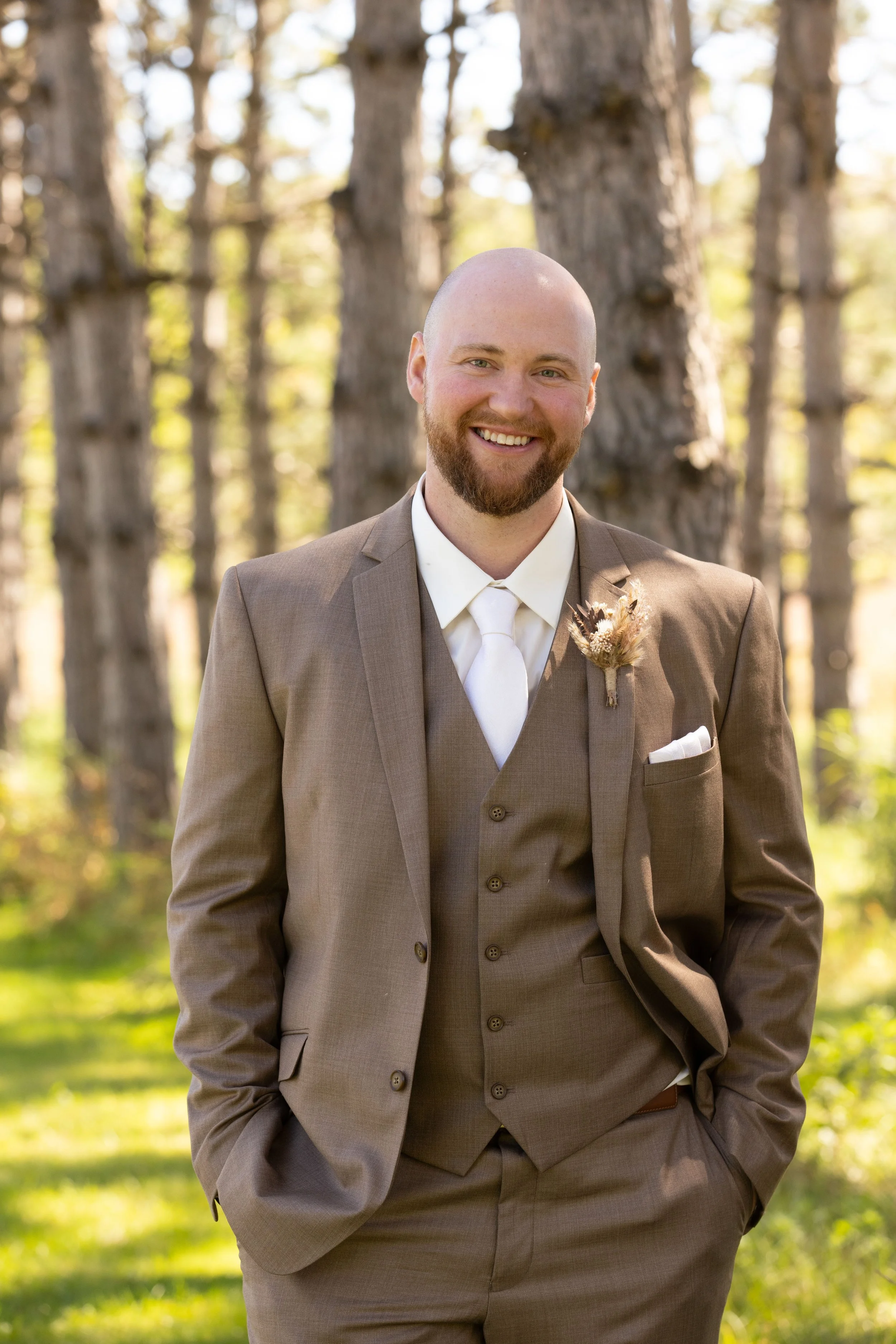 A groom in a brown suit with a boutonniere stands outdoors in a forest, smiling with hands in his pockets.