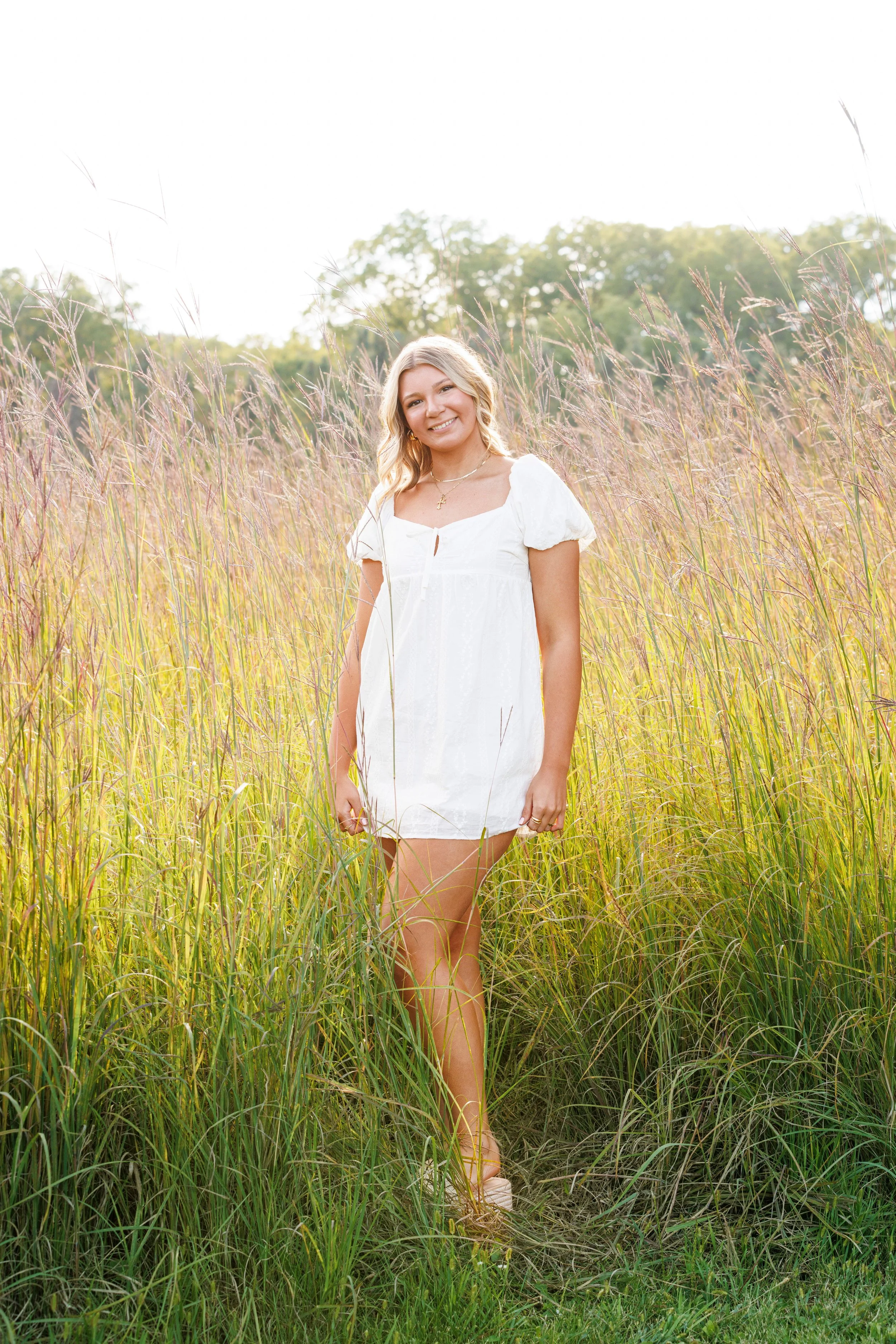 A young woman with blonde hair in loose waves, wearing a short white dress and sandals, standing in a tall grassy field during daytime, smiling at the camera.