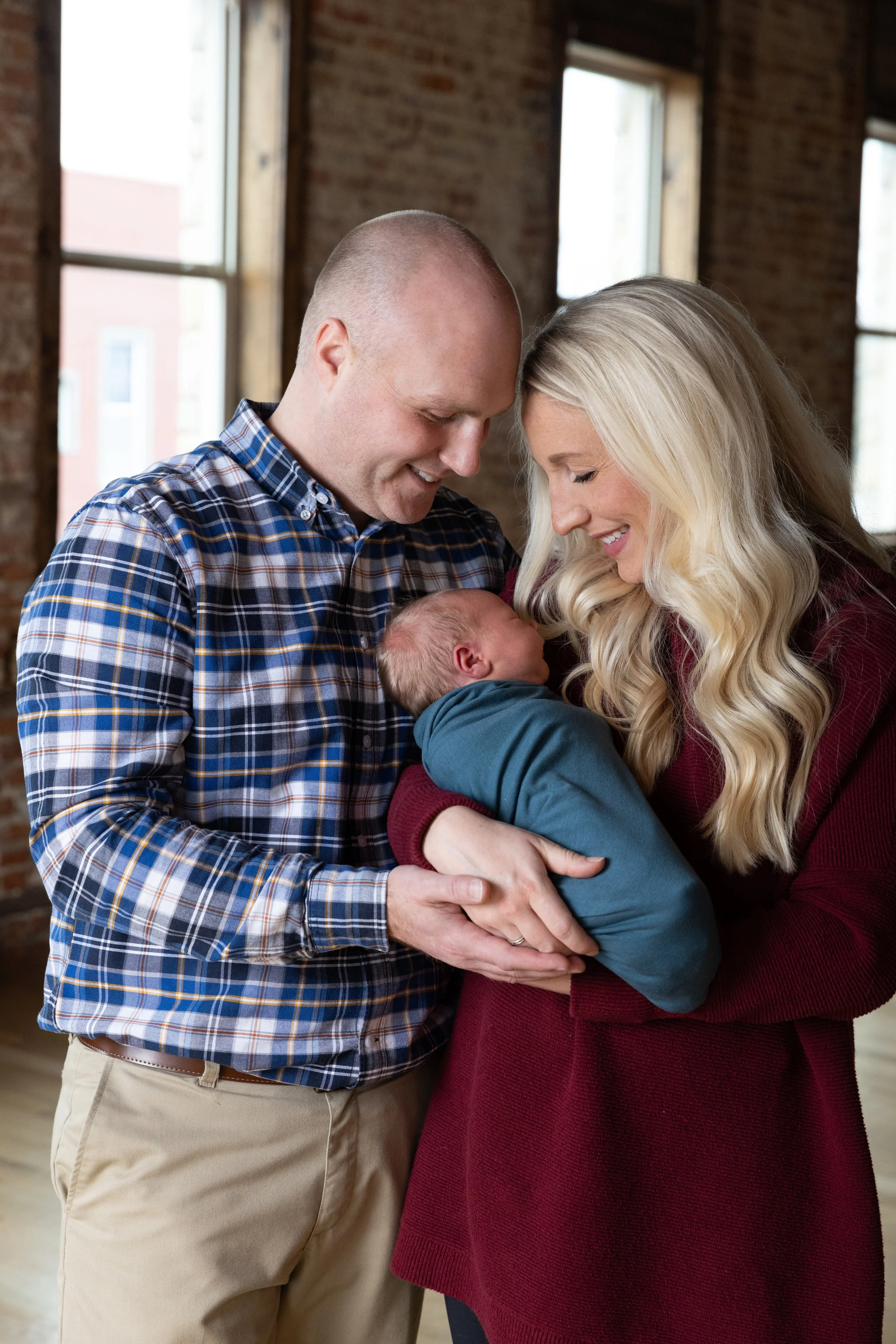 A couple holding a newborn baby inside a brick-walled room with large windows, with both adults smiling and looking affectionately at the infant.