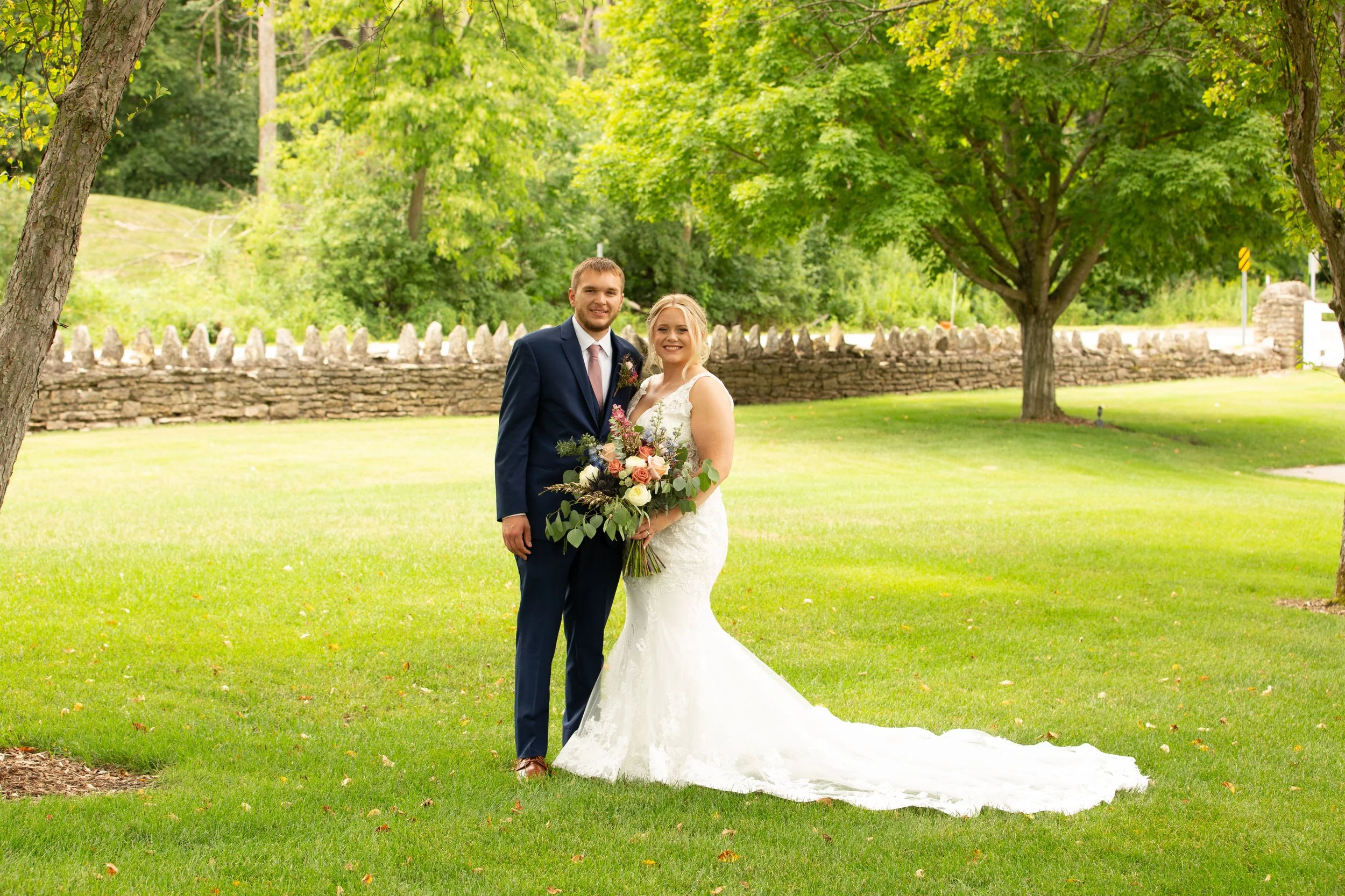 A bride and groom standing together outdoors on a grassy lawn, with trees and a stone wall in the background. The bride is wearing a white wedding gown and holding a bouquet. The groom is dressed in a navy suit with a tie.