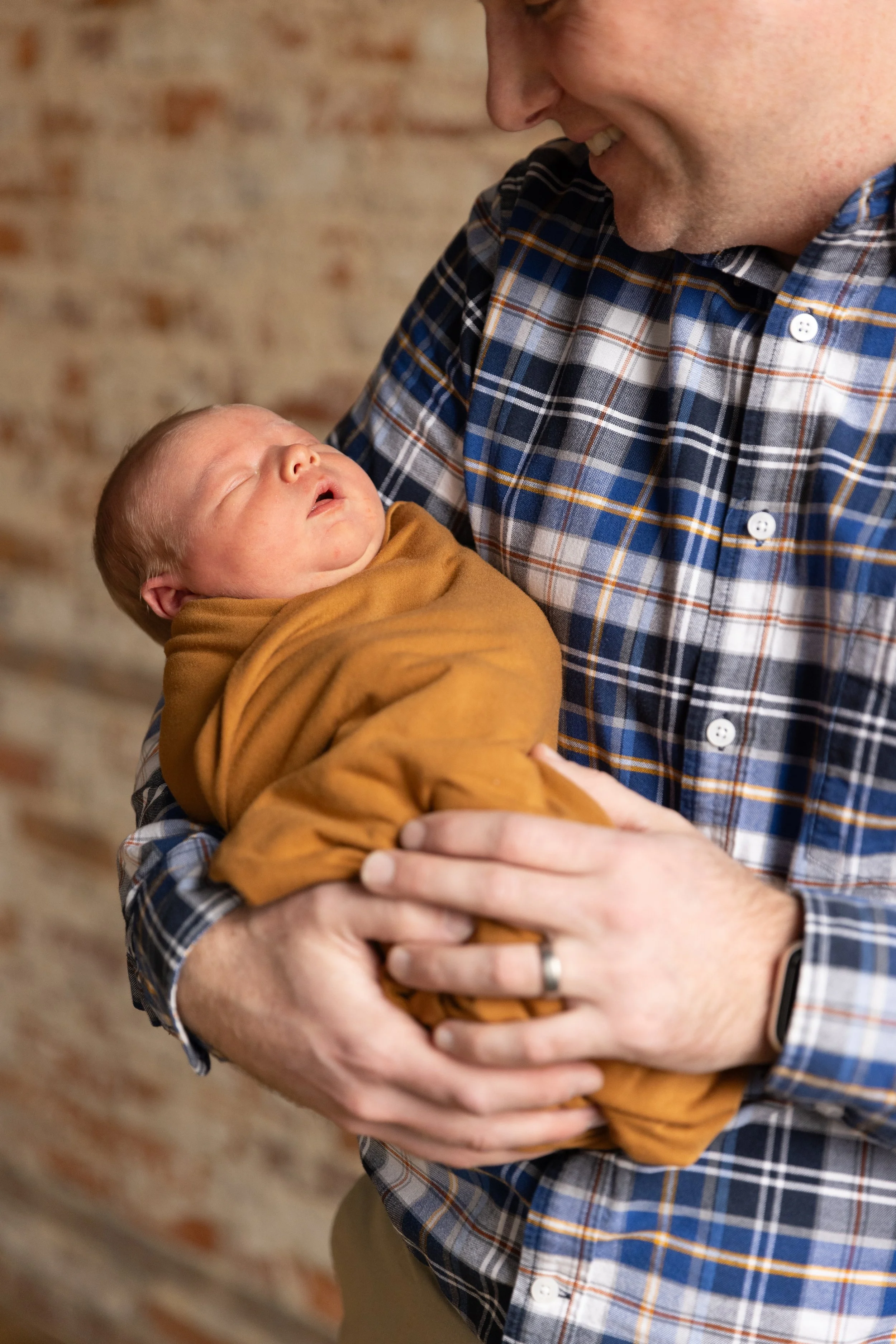 A man holding a sleeping newborn baby wrapped in a brown blanket, smiling and looking down at the baby.